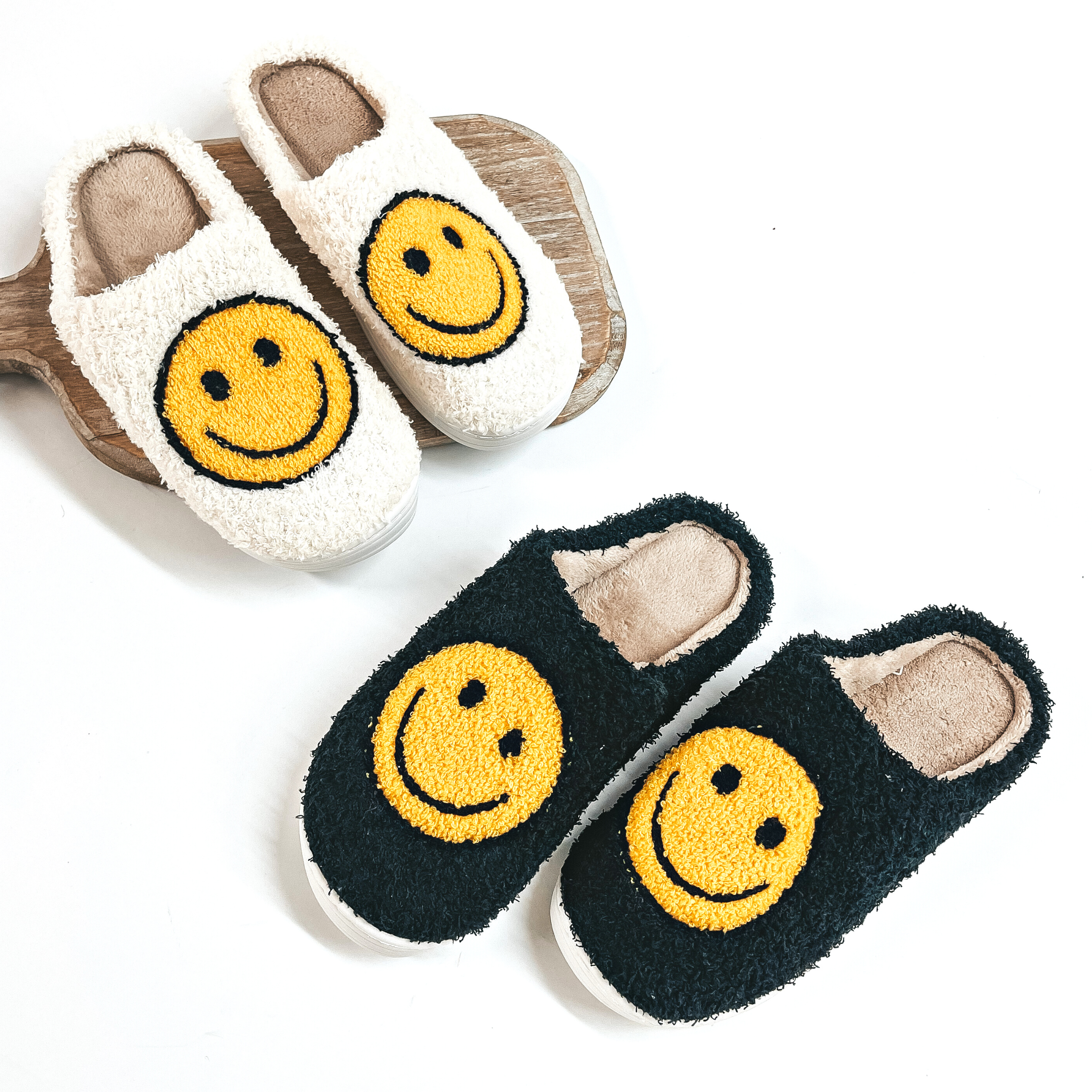 There are two pairs of sherpa happy face slippers in ivory and black, both have  brown inner soles. The ivory slippers are placed on a wooden slab and the  black pair is on a white background.