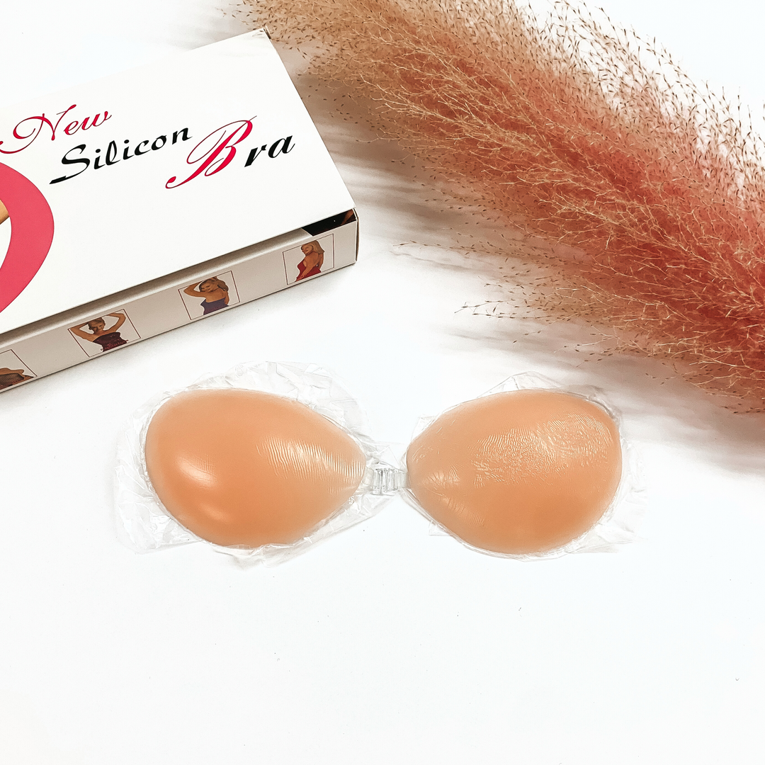 There is a nude color silicone clip on bra, there is a a white box in the back that shows the packaging. All items are in a white background with a pink plant in the side as decor.