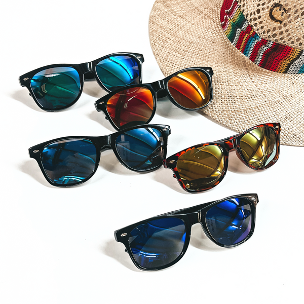 There are five pairs of sunglasses with different colored lenses and frames. These sunglasses are taken on a white background and a straw hat with a colorful hat band. 