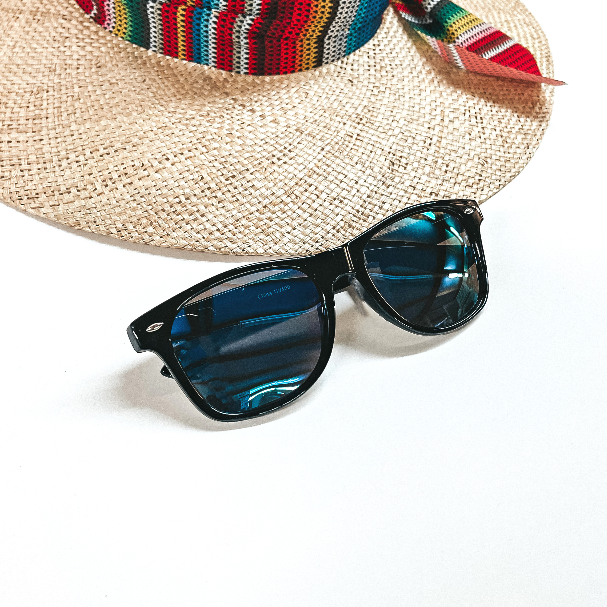 These are black framed sunglasses with dark blue lenses and silver detailing. These sunglasses are placed on a white background with a straw hat in the back as decor.