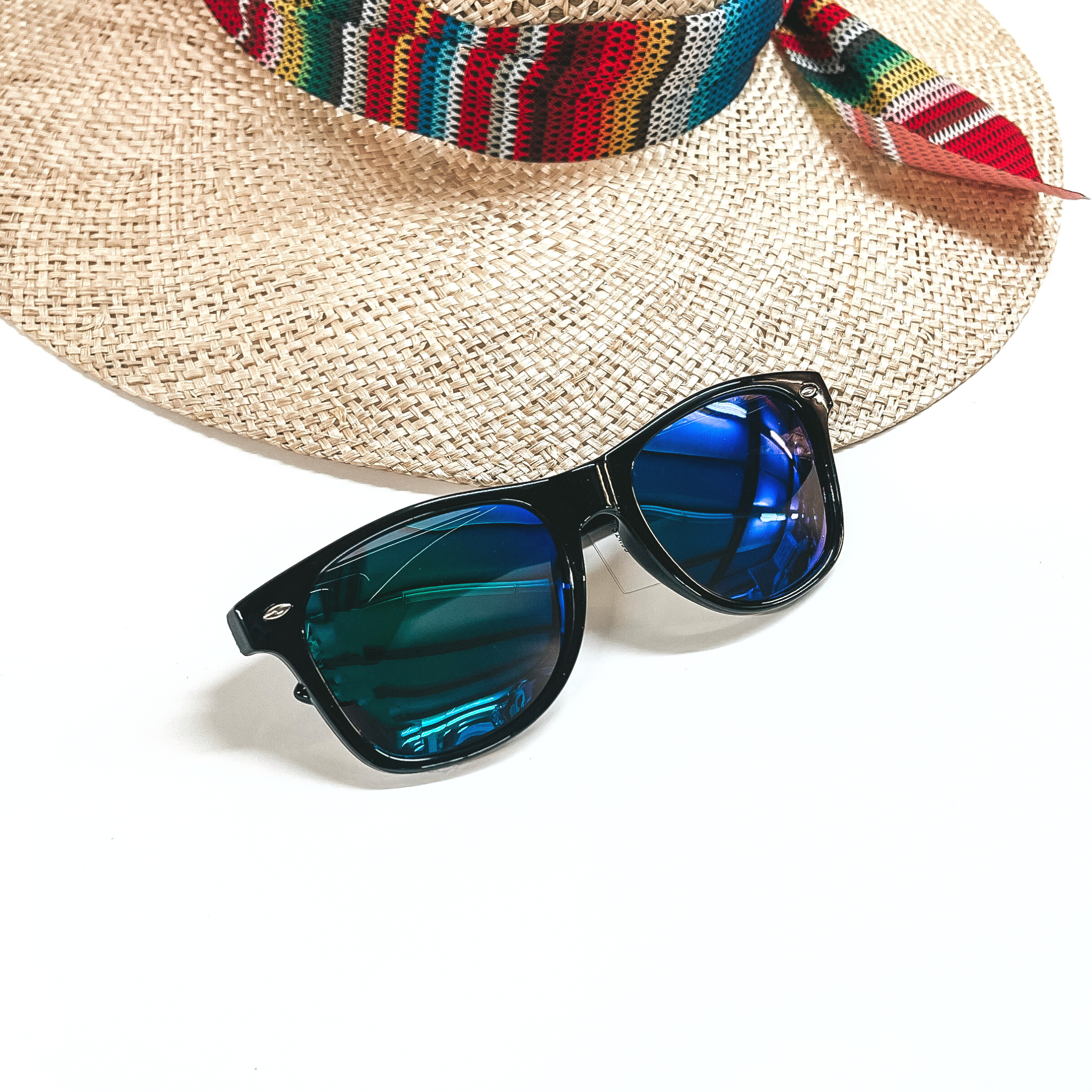 These are black framed sunglasses with blue/green lenses and silver detailing. These sunglasses are taken on a white background and a straw hat with a colorful hat band in the back as decor.