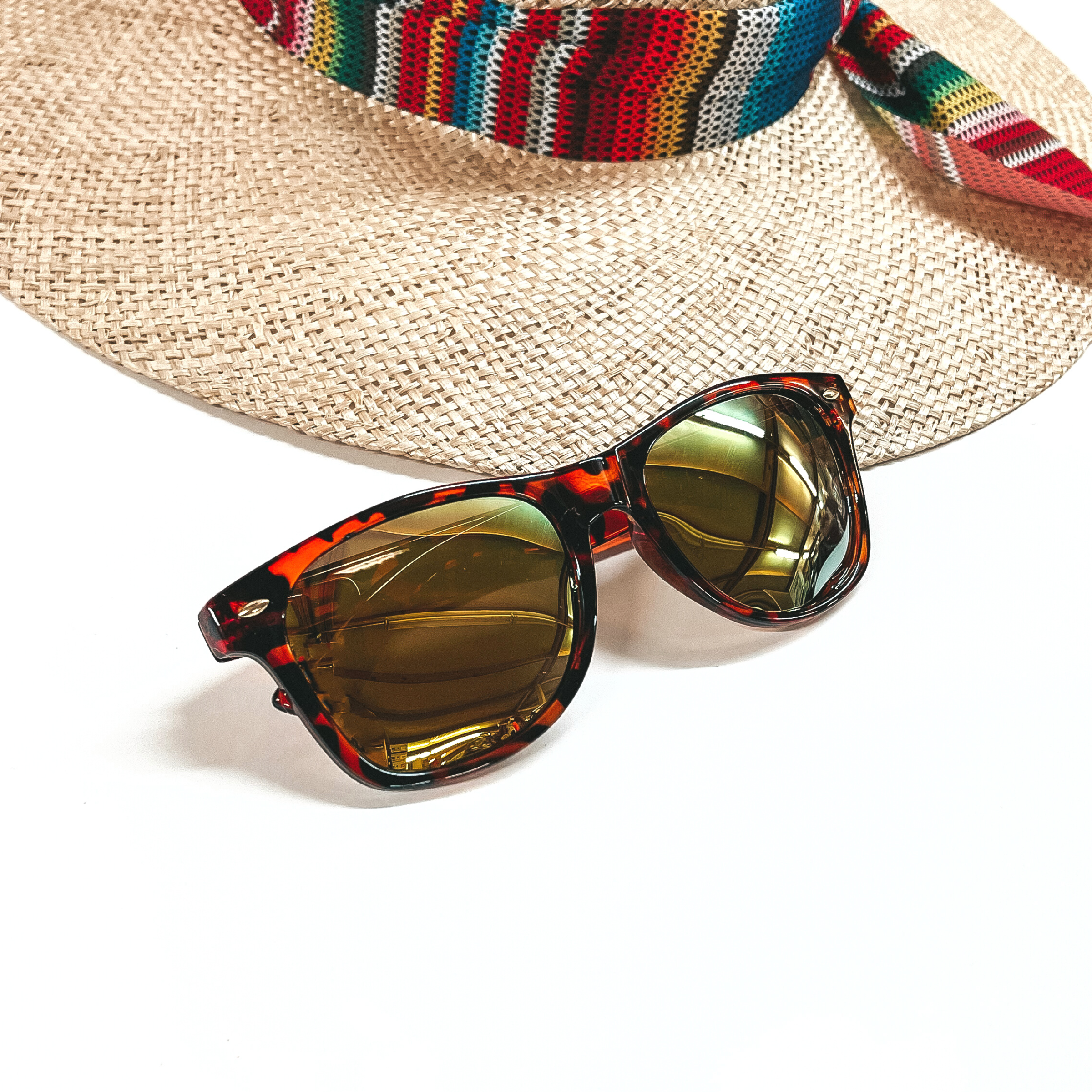 These are tortouise print frames with a yellow/green lense and silver detailing. These sunglasses are placed on a white background and a straw hat with a colorful hat band in the back as decor.