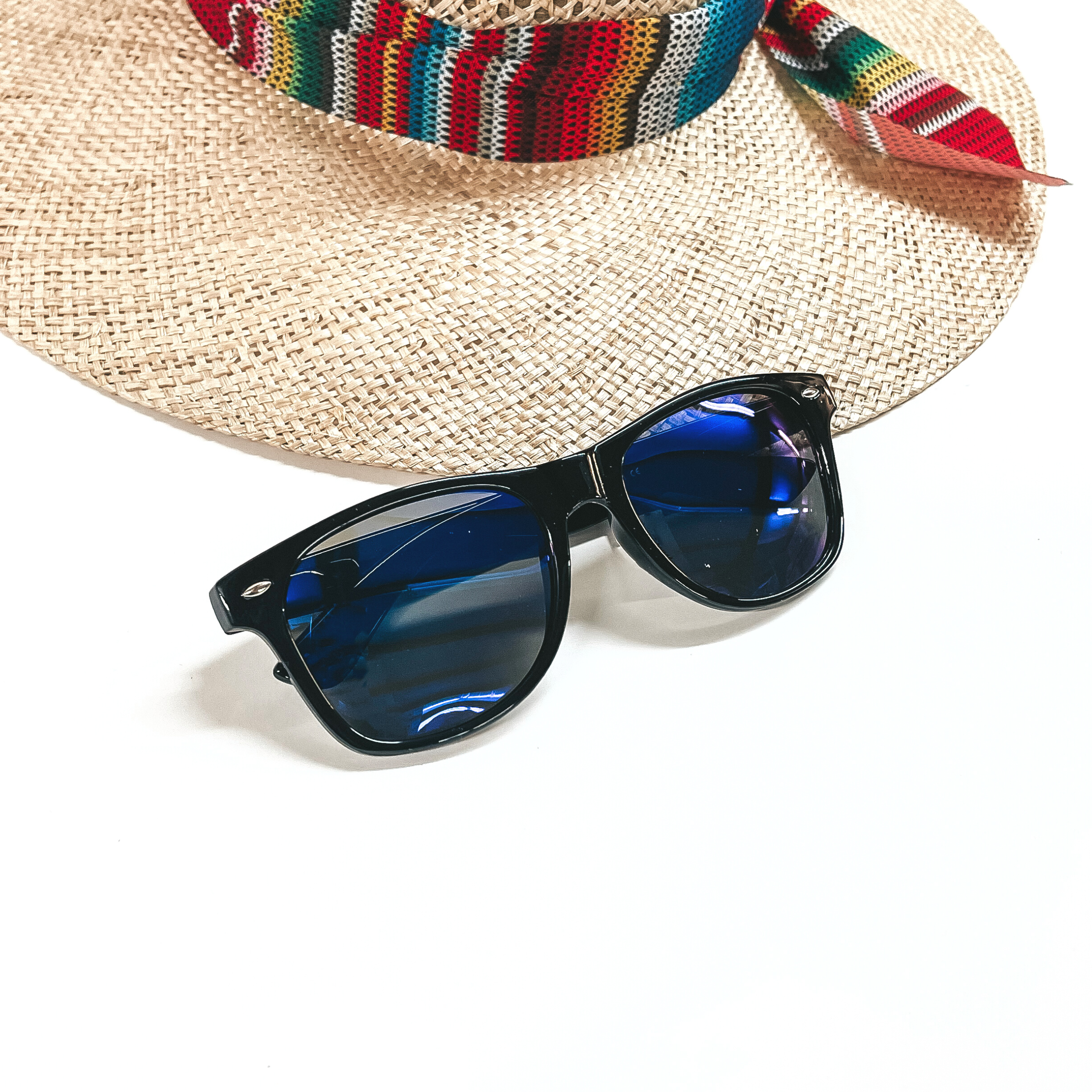 These are black framed sunglasses with blue/dark purple lenses and silver detailing. These sunglasses are placed on a white background and a straw hat with a colorful hat band in the back as decor.
