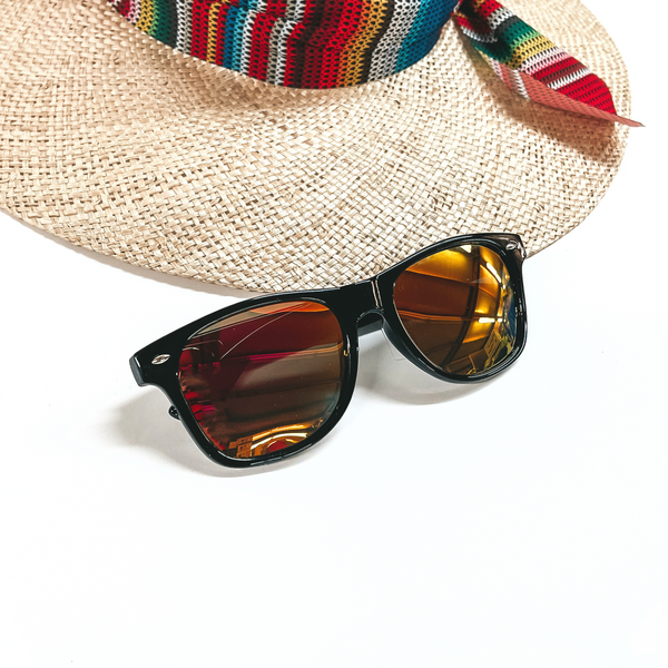 These are black framed sunglasses with an orange/yellow lense and silver detailing. These sunglasses are taken on a white background and a straw hat with a colorful hat band in the back as decor.