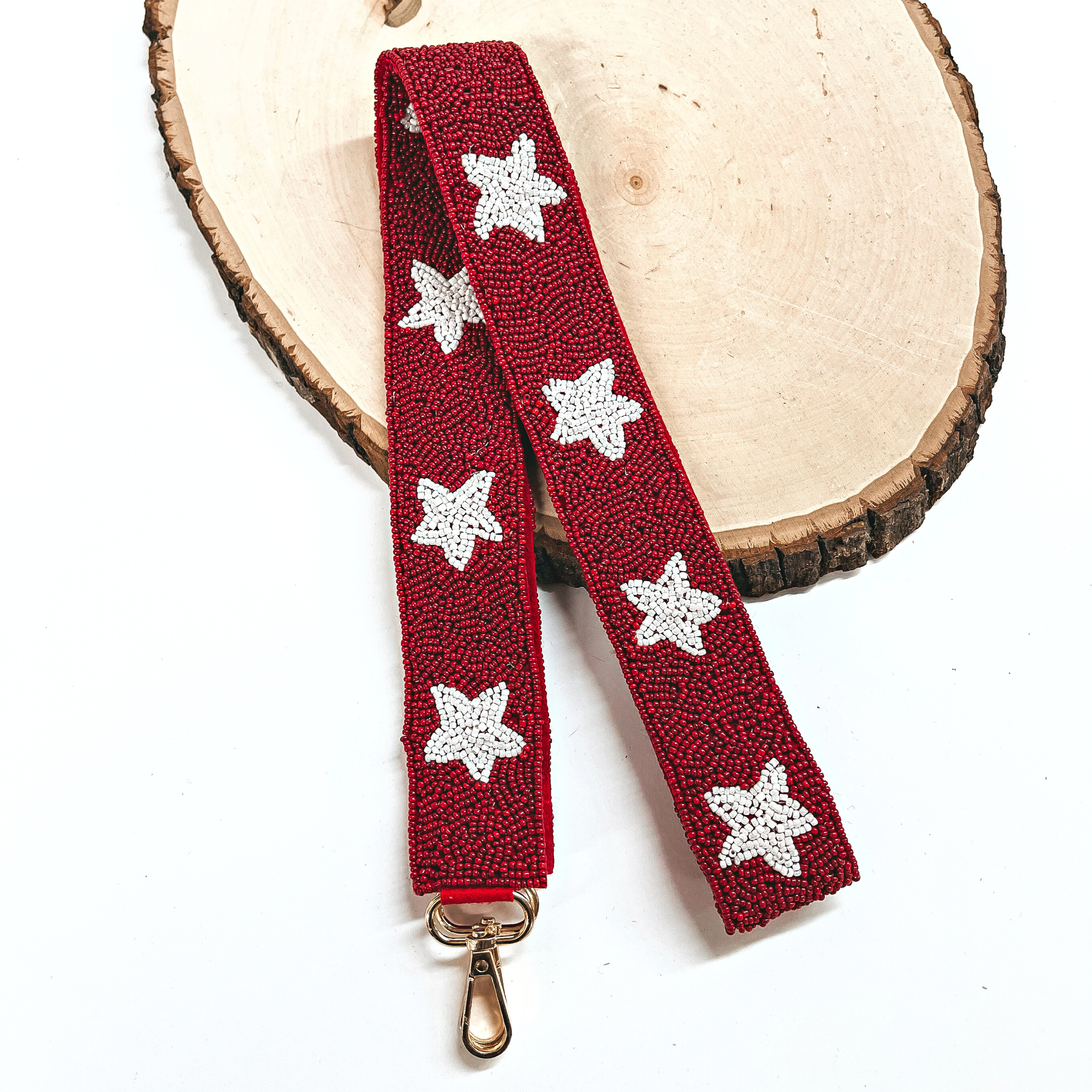 This is a maroon/dark red beaded purse strap with white beaded stars all along and a gold clasp. This purse strap is taken on a slab of wood and on a white background.