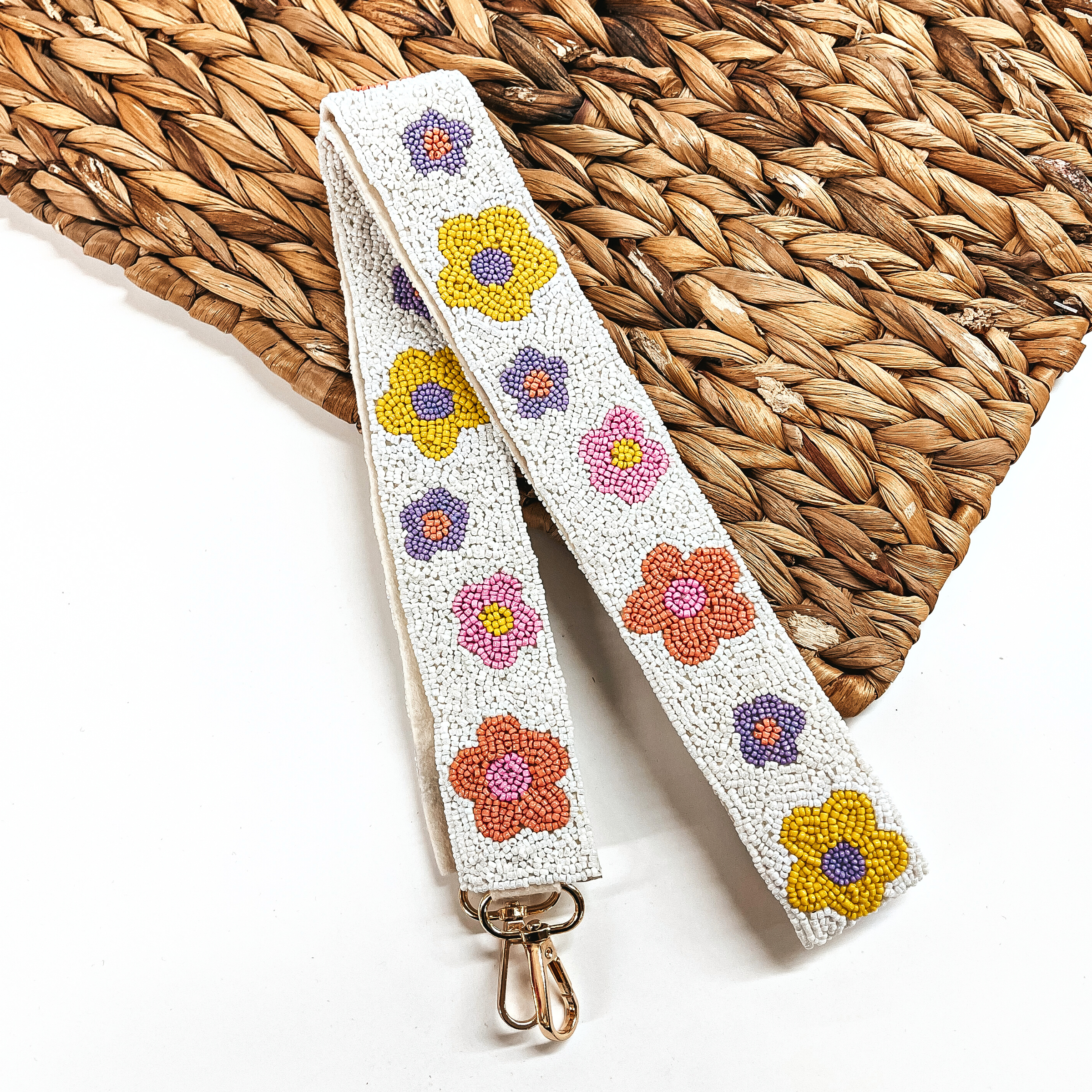 This is a white beaded purse strap with pastel colored flowers all along in different sizes, with a gold clasp. This purse strap is taken on a white background and a brown woven slate.