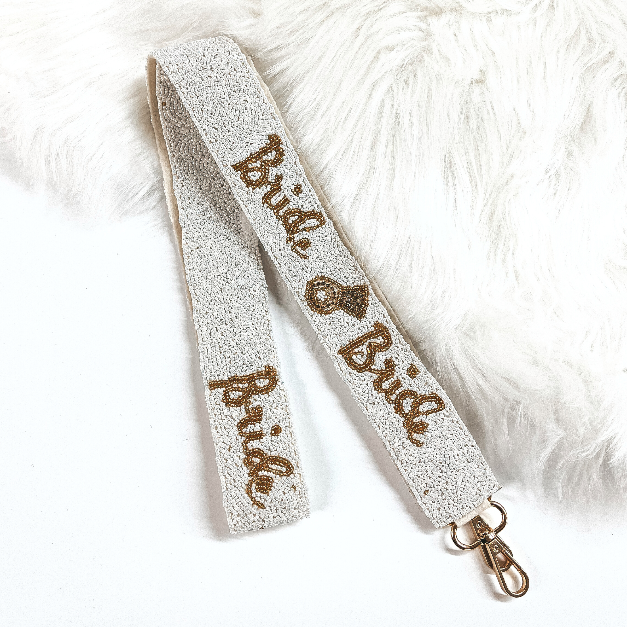 This is a white beaded purse strap that say's 'Bride' with an engagement ring in gold. This purse strap is taken on white background and on white fur.