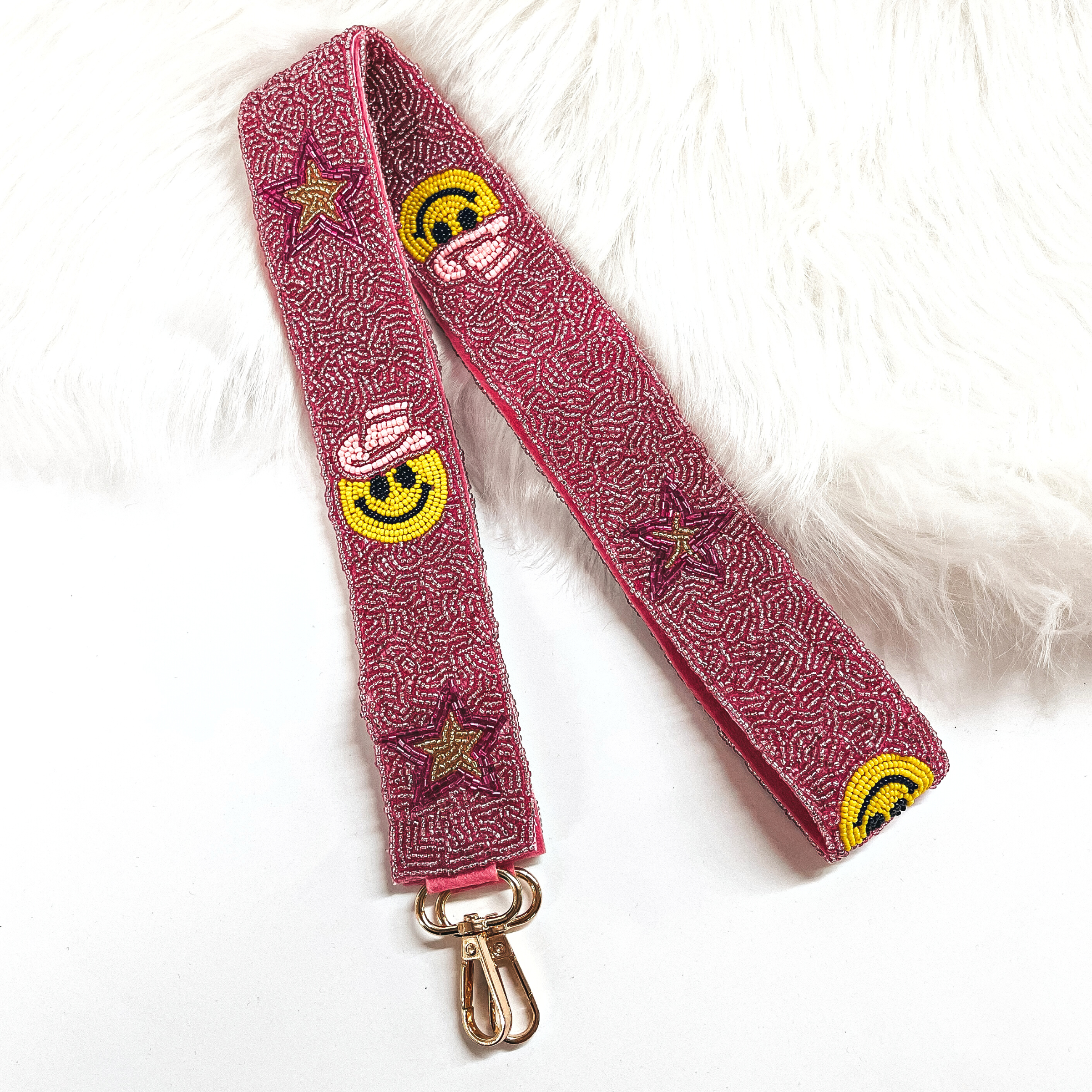 This is a pink beaded purse straps with pink and gold beaded stars, and a yellow happy face with a pink cowboy hat. This purse strap is taken on a white background and on white fur.