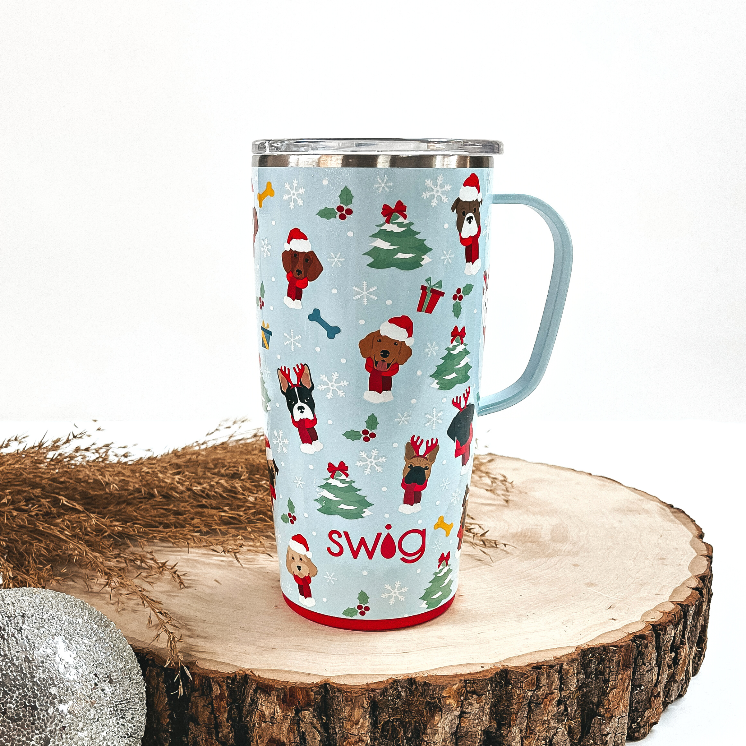 This is a light blue cup with a handle, a red top and bottom, with different kinds  of dogs, christmas tree's, snowflakes, mistletoes, and presents all over.  This cup is taken on top of a slab of wood and on a white background, with  a silver ornament and brown plant in the back as decor.