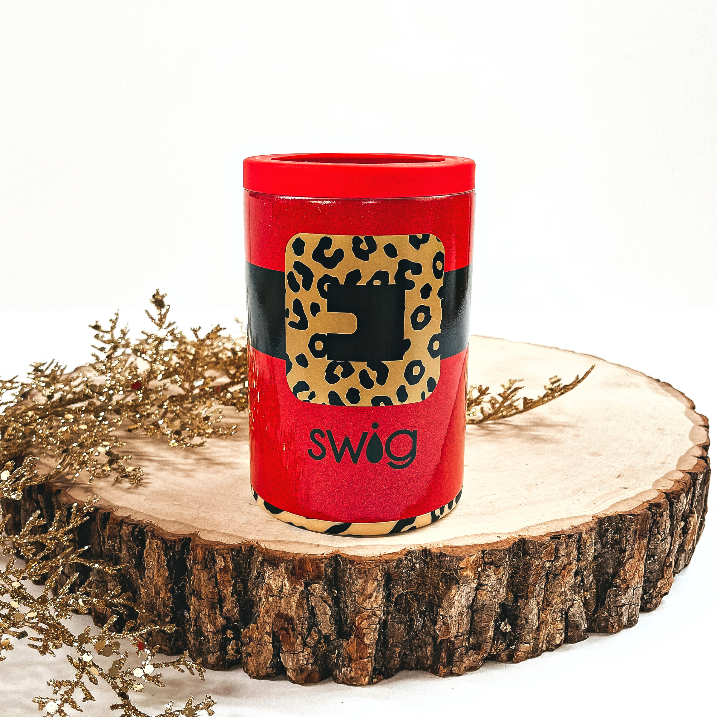This is a red cup with a black thick line all around and a gold/black  leopard print buckle. It has a red top and a leopard print bottom. This cup is taken on top of a slab of wood and on a white background, with  a gold sparkly plant in the side as decor.