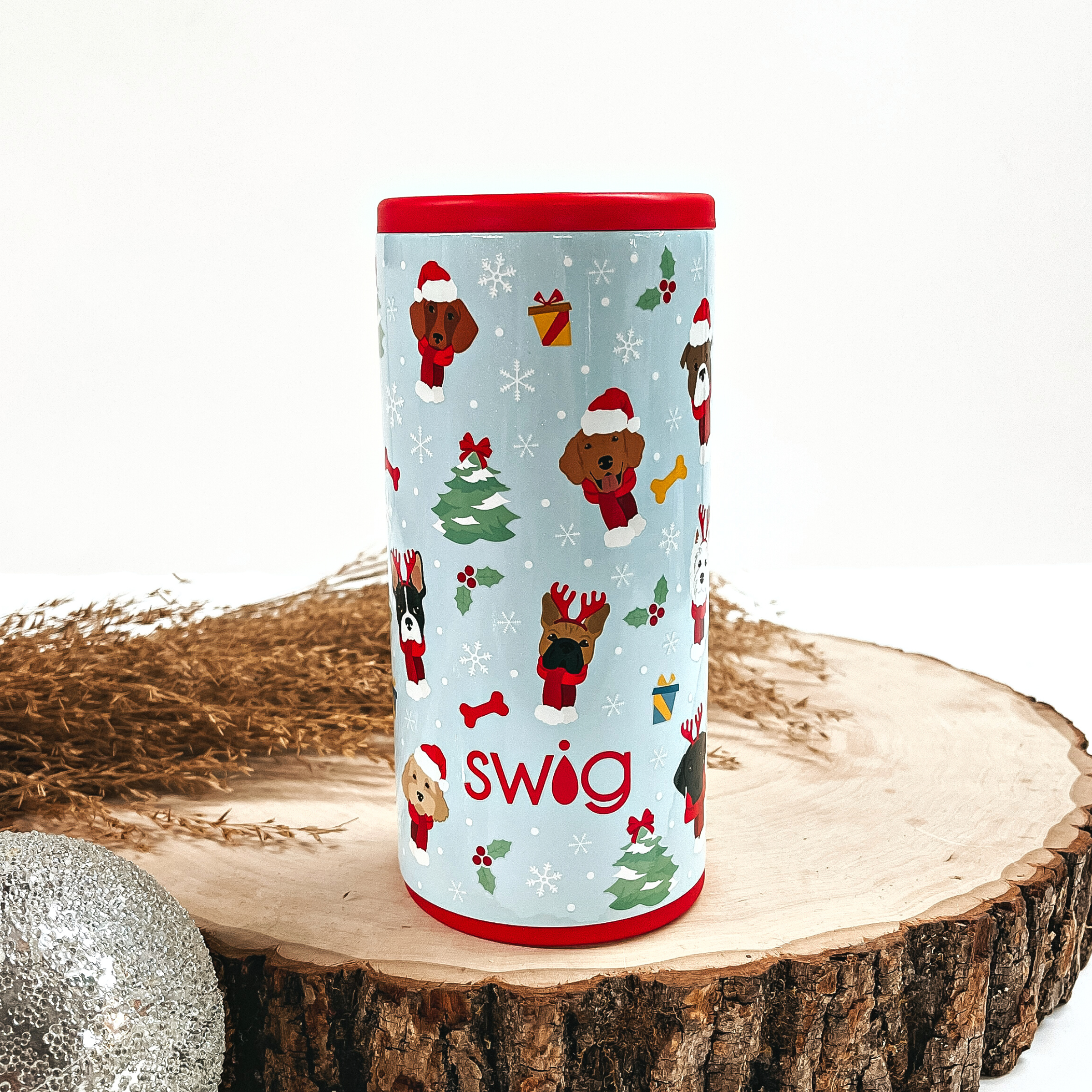 This is a light blue cup with a red top and bottom, with different kinds  of dogs, christmas tree's, snowflakes, mistletoes, and presents all over.  This cup is taken on top of a slab of wood and on a white background, with  a silver ornament and brown plant in the back as decor.