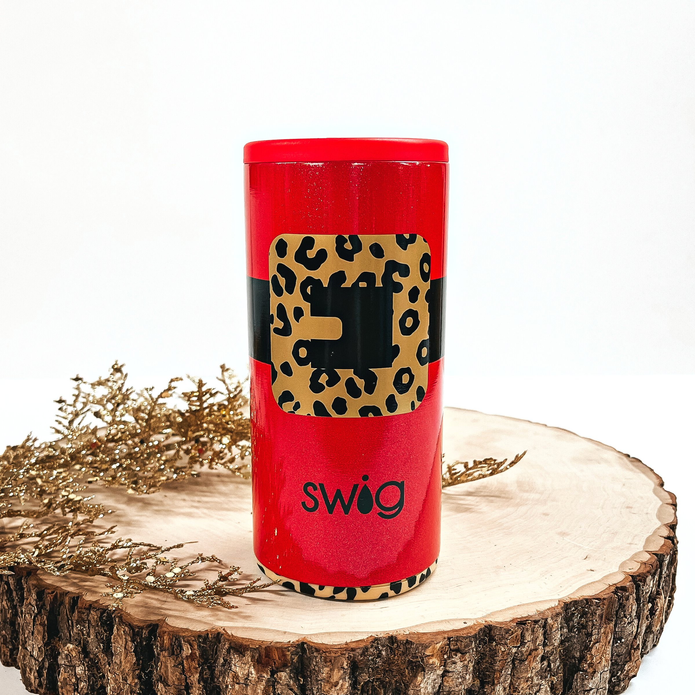 This is a red cup with a black thick line all around and a gold/black  leopard print buckle. It has a red top and a leopard print bottom. This cup is taken on top of a slab of wood and on a white background, with  a gold sparkly plant in the side as decor.