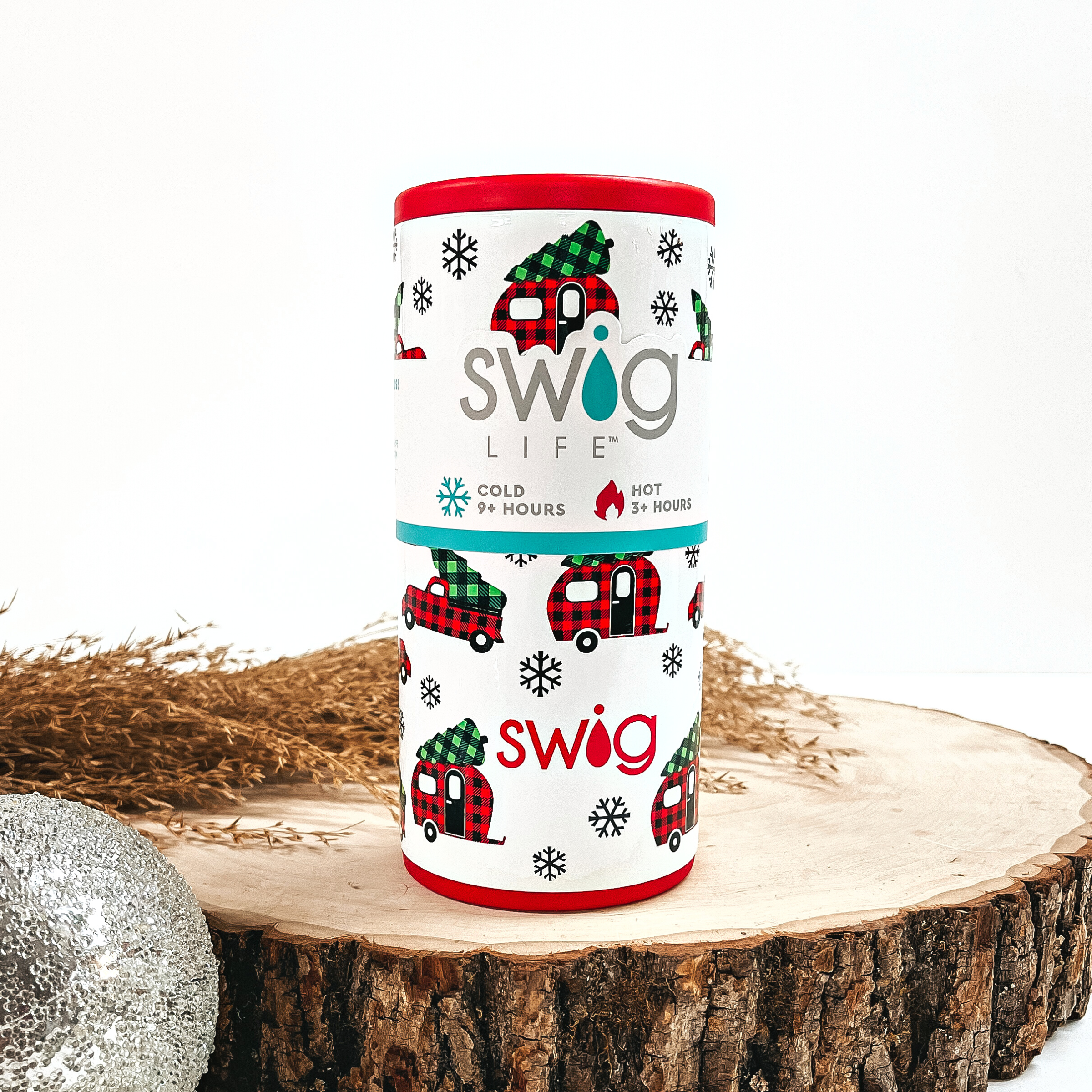 This is a white cup with a red top and bottom, with red buffalo plaid  trucks and campers, green buffalo plaid christmas tree's, and black  snowflakes.  This cup is taken on top of a slab of wood and on a white background, with  a silver ornament and brown plant in the back as decor.