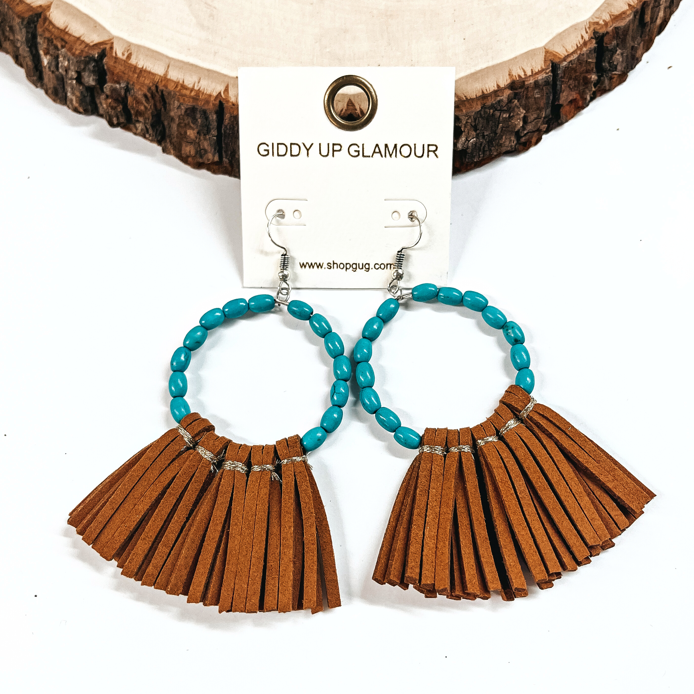 These are oval turquoise beaded hoops with a faux leather tassel fringe  in brown. These earrings are placed on an ivory Giddy Up Glamour  earring card. These earrings are taken leaning up against a  slab of wood and on a white background.