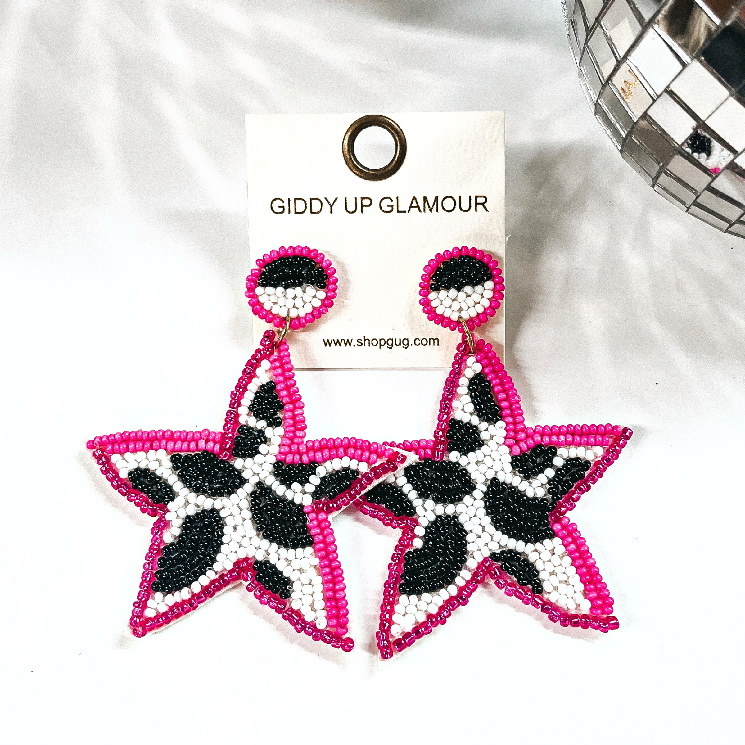 These are beaded star shape earrings in pink, black, and white. The center  is in black and white cow print, and pink beads all around. The post  back is half black and white, with pink beads all around as well. These  earrings are placed on an ivory Giddy Up Glamour earring card. These earrings  are taken on a white background with a disco ball in the side as decor.