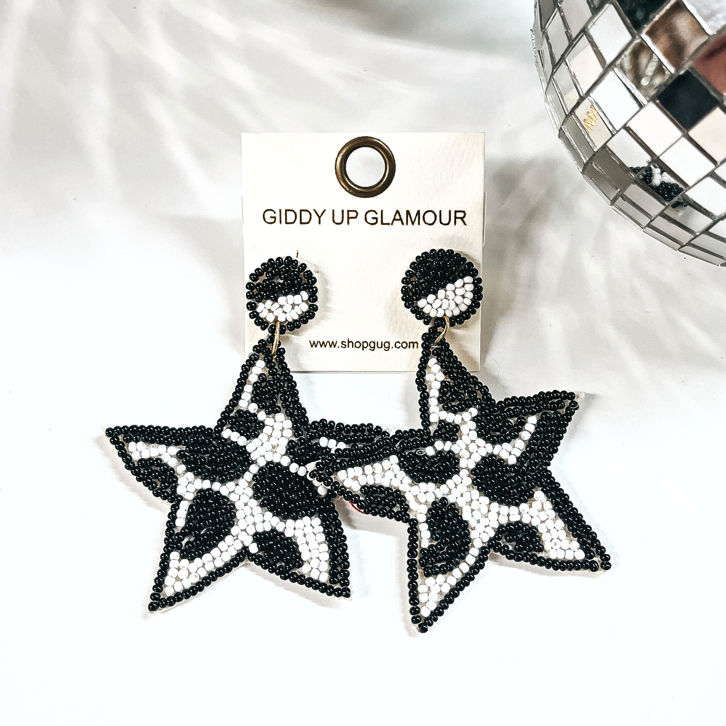 These are beaded star shape earrings in black and white. The center  is in black and white cow print, and black beads all around. The post  back is half black and white, with black beads all around as well. These  earrings are placed on an ivory Giddy Up Glamour earring card. These earrings  are taken on a white background with a disco ball in the side as decor.
