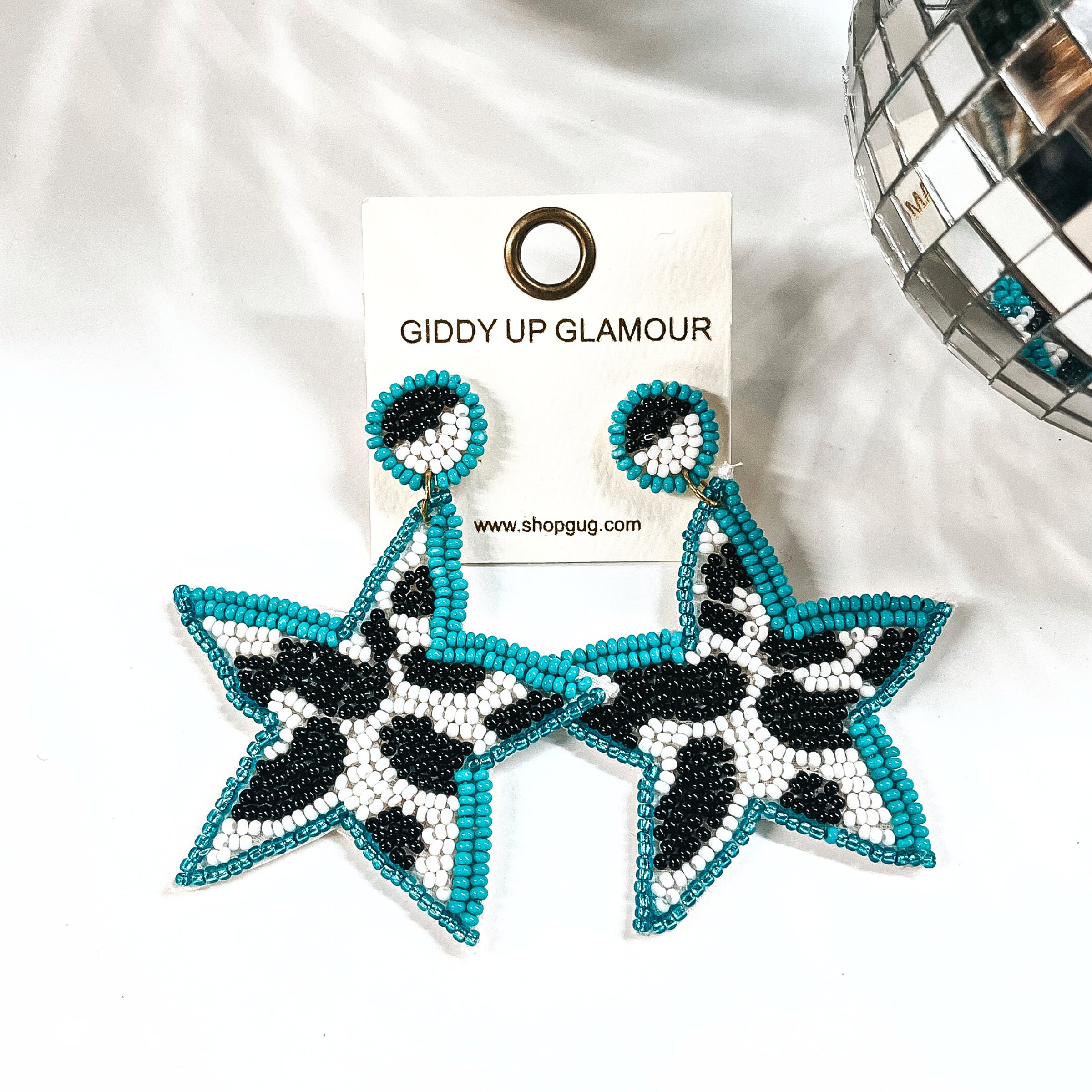These are beaded star shape earrings in turquoise, black, and white. The center  is in black and white cow print, and turquoise beads all around. The post  back is half black and white, with turquoise beads all around as well. These  earrings are placed on an ivory Giddy Up Glamour earring card. These earrings  are taken on a white background with a disco ball in the side as decor.