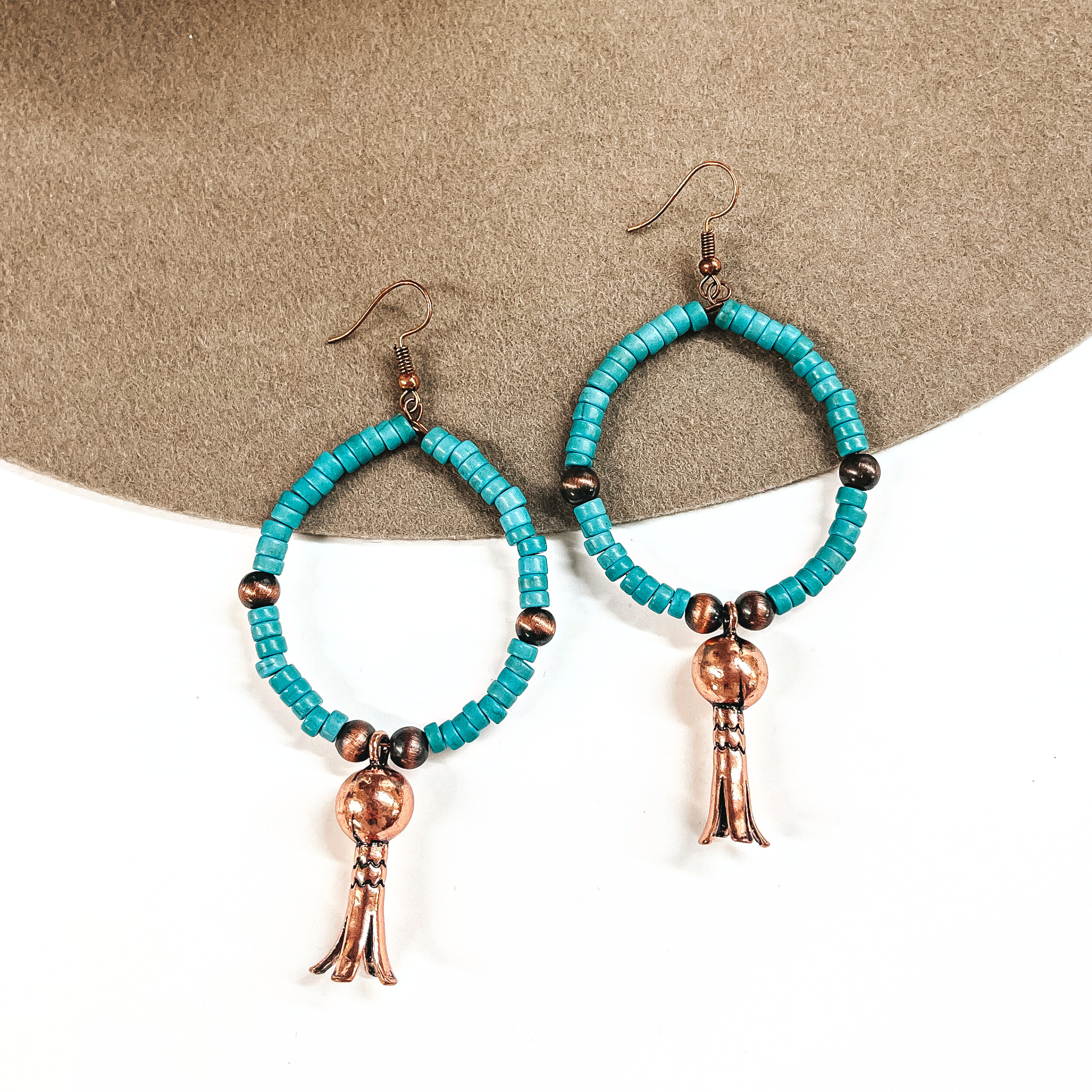 These are turquoise beaded hoop earrings with copper bead spacers, and a copper squash  blossom drop. These earrings are taken on a brown felt hat brim and on a  white background.