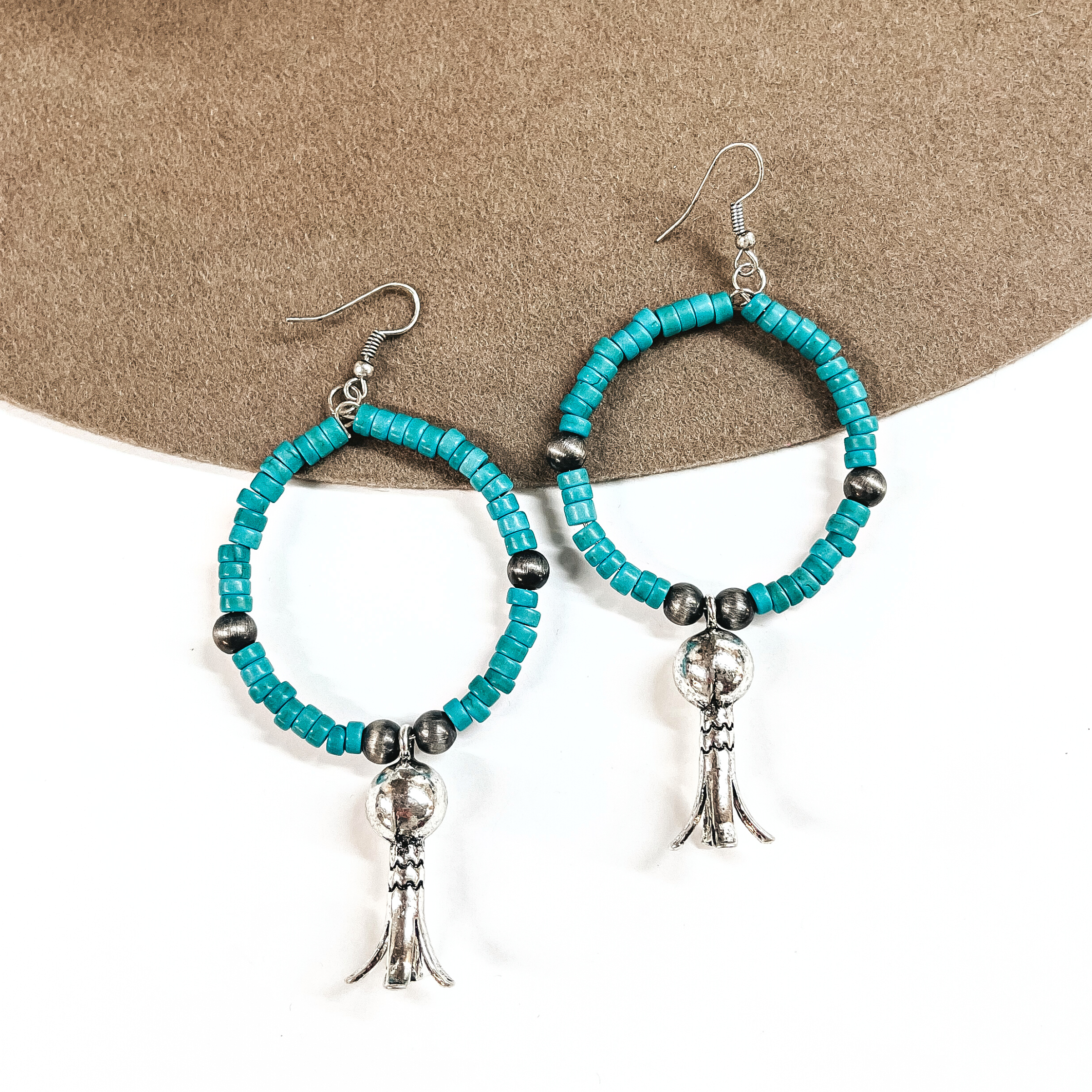These are turquoise beaded hoop earrings with silver bead spacers, and a silver squash  blossom drop. These earrings are taken on a brown felt hat brim and on a  white background.