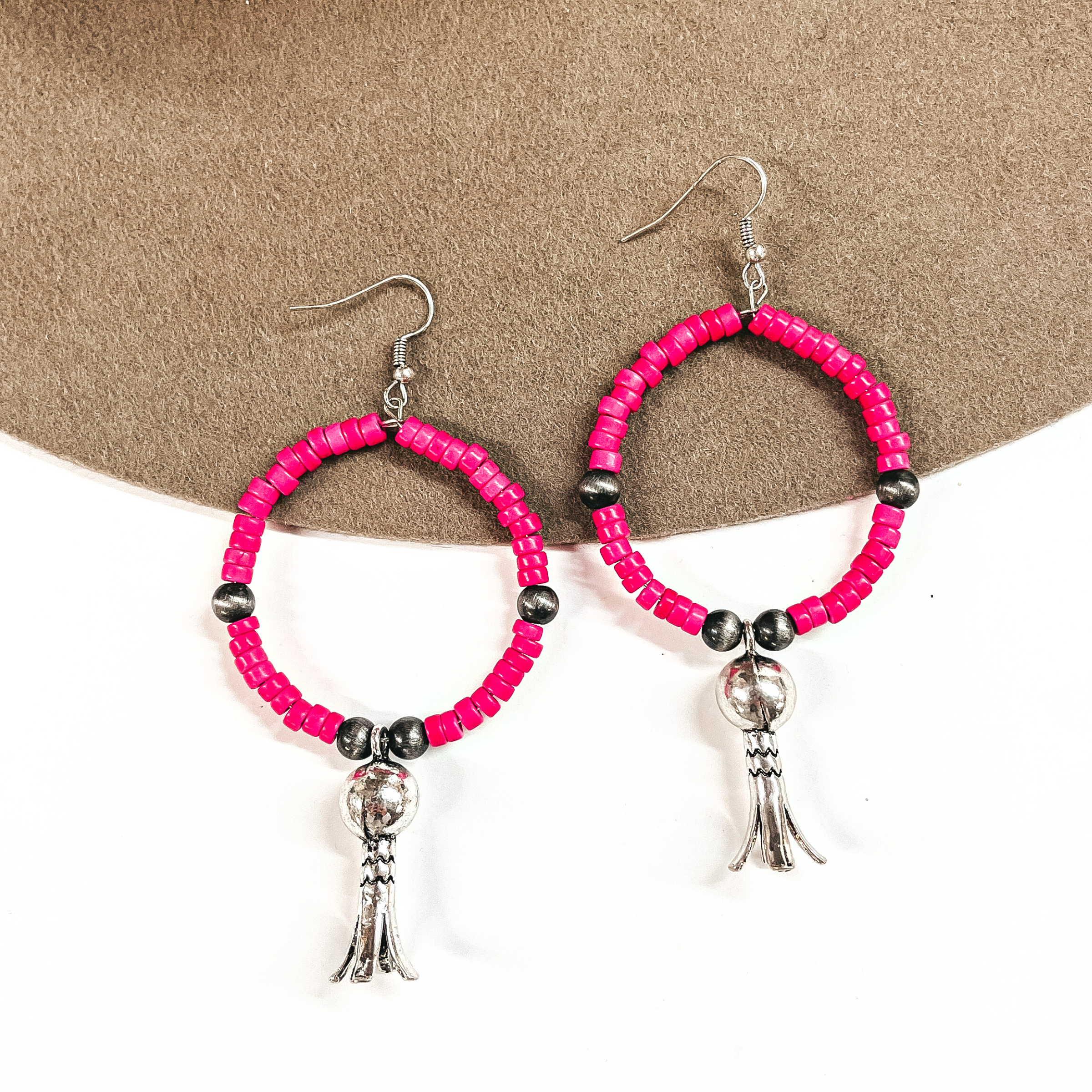 These are pink beaded hoop earrings with silver bead spacers, and a silver squash  blossom drop. These earrings are taken on a brown felt hat brim and on a  white background.