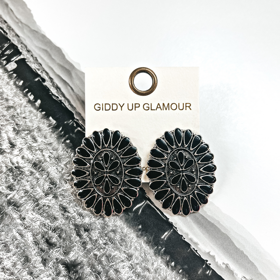 These are oval concho earrings in a silver settings with black stones all  around and in the center. These earrings are placed on an ivory Giddy Up  Glamour earrings card. These earrings are taken on a black and white western  page.