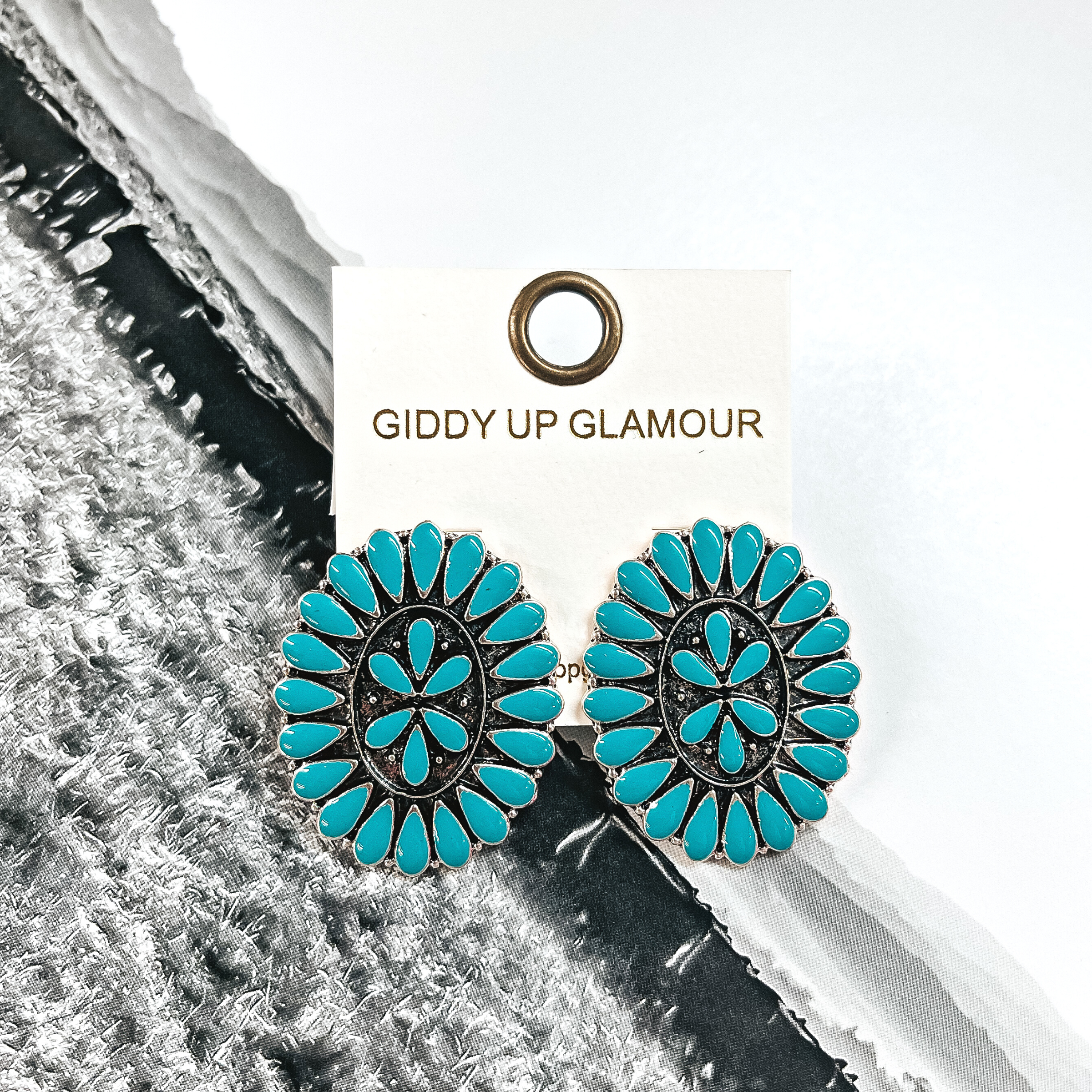 These are oval concho earrings in a silver setting with turquoise stones all  around and in the center. These earrings are placed on an ivory Giddy Up  Glamour earrings card. These earrings are taken on a black and white western  page.