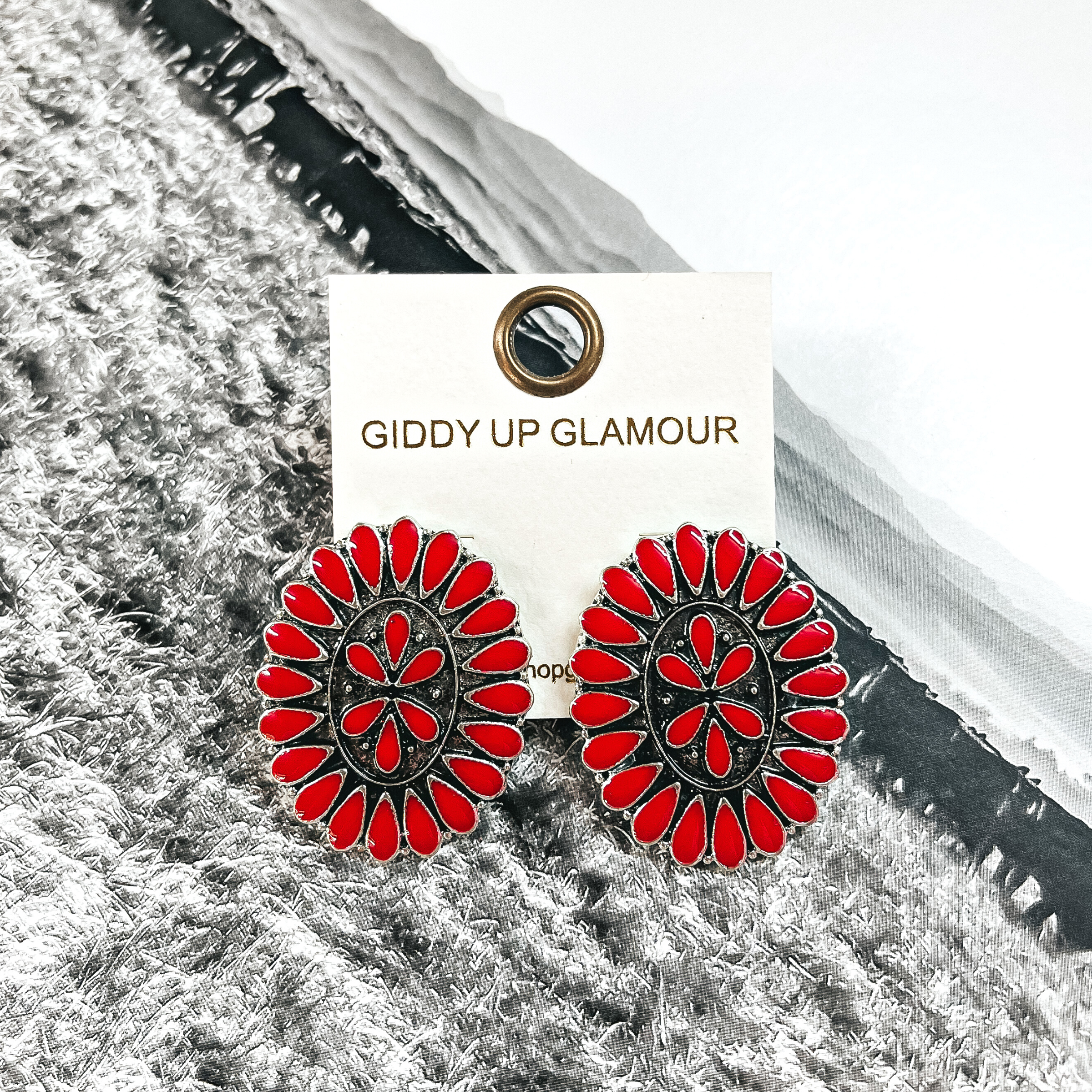 These are oval concho earrings in a silver setting with red stones all  around and in the center. These earrings are placed on an ivory Giddy Up  Glamour earrings card. These earrings are taken on a black and white western  page.