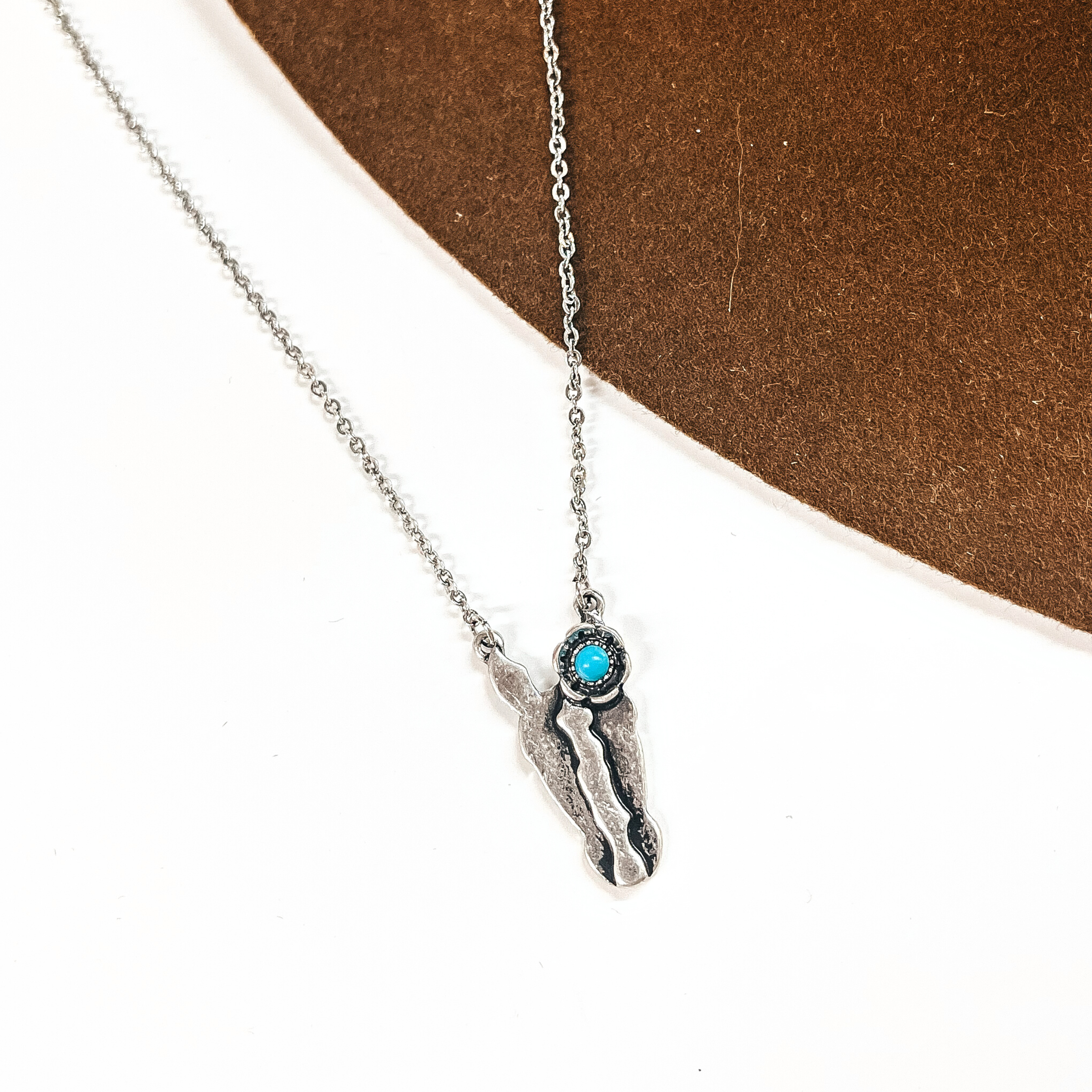 This is a silver chain necklace with a horse head pendant, has a small turquoise stone on the ear. This necklace is taken on a white background and on a dark brown felt hat brim.