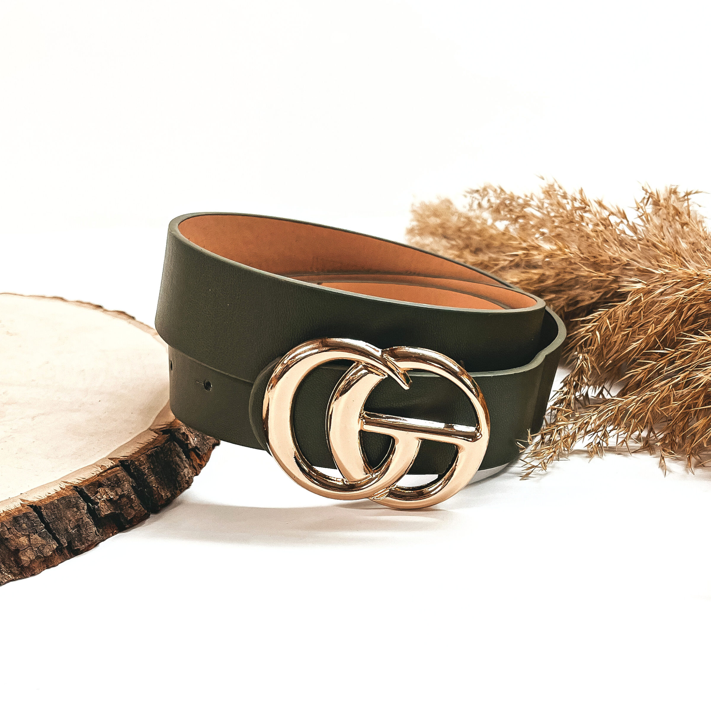 This is a olive green rolled up belt with a gold buckle in the center. This belt  is placed on a slab of wood and on a white background, there is a brown plant  in the back as decor.