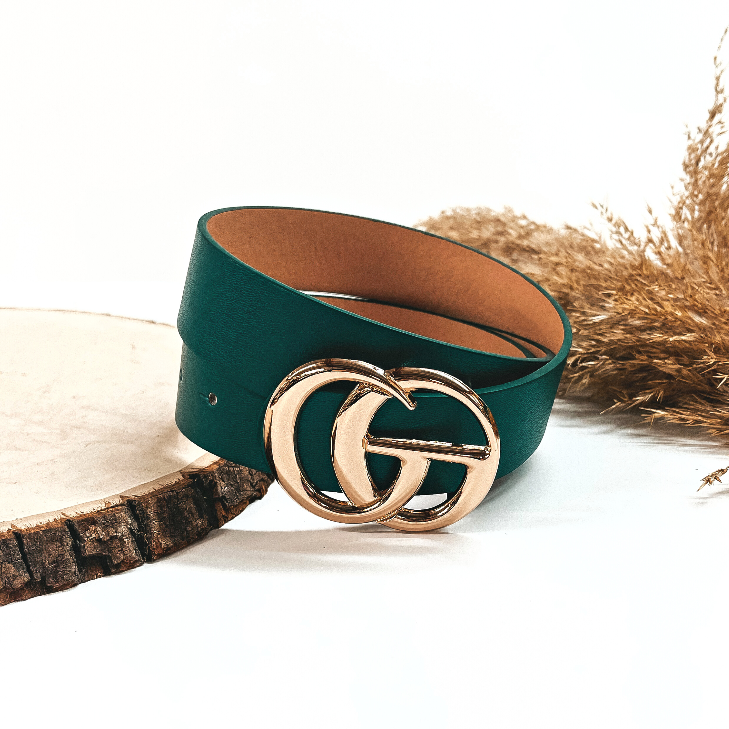 This is a dark green rolled up belt with a gold buckle in the center. This belt  is placed on a slab of wood and on a white background, there is a brown plant  in the back as decor.