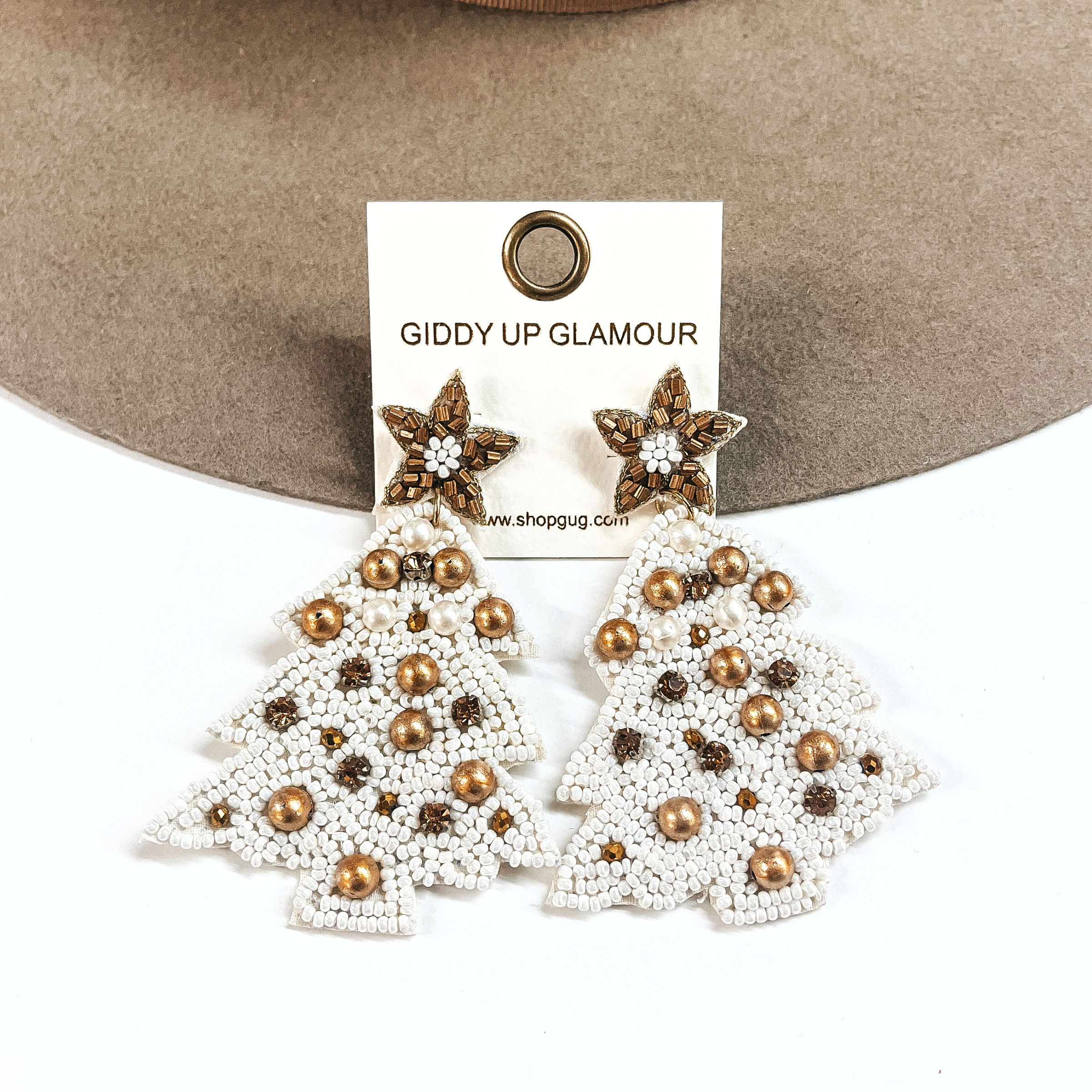 This is an ivory/white beaded christmas tree earrings with a gold beaded postback. There are matte gold beads all over and topaz colored crystals as well. These earrings are taken leaning against a light brown felt hat and on a white background.