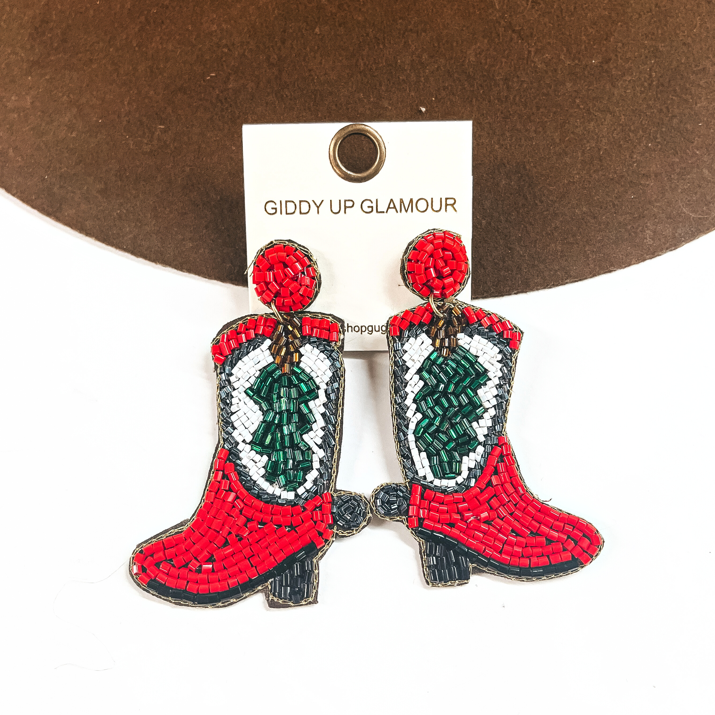 These are beaded Christmas boot earrings in red, white, black, green, brown, and gray. It has a red postback with a boot drop in red, the calf part of the boot has a beaded tree in a white background, and has a black sole. These earrings are taken leaning against a dark brown felt hat and on a white background.