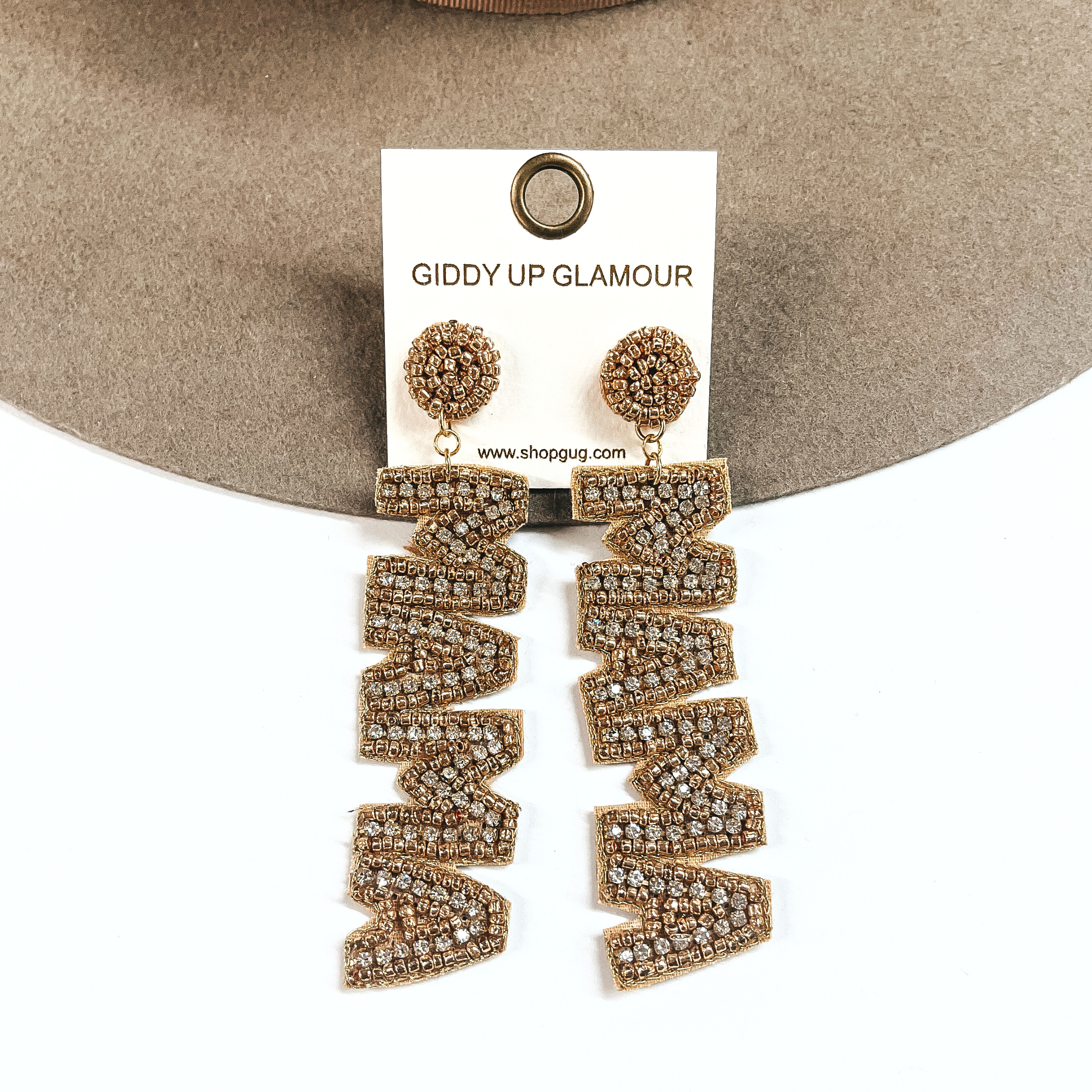 These are gold beaded and clear crystal earrings with a drop that says, MAMA. These earrings are taken on a light brown felt hat brim and on a white background.