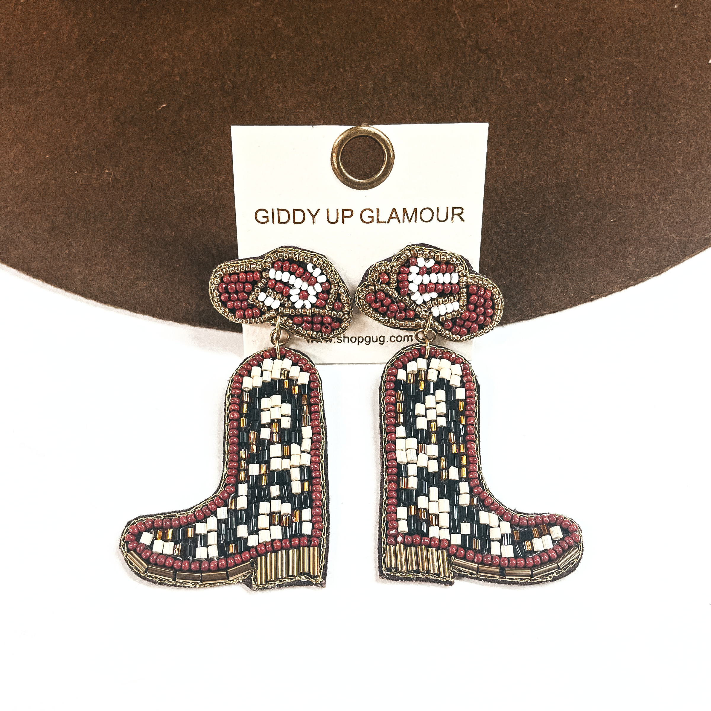 These are hat and boot beaded earrings in brown, white, gold, and black. The hat is the postback in brown and white with gold beads all around. There is a boot drop with a gold beaded sole. These earrings are leaning up against a dark brown felt hat brim and on a white background.