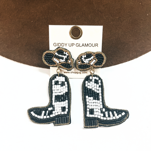 These are hat and boot beaded earrings in white and black. The postback is a hat with gold beads all around and with a boot drop. These earrings are taken leaning up against a dark brown felt hat brim and on a white background.