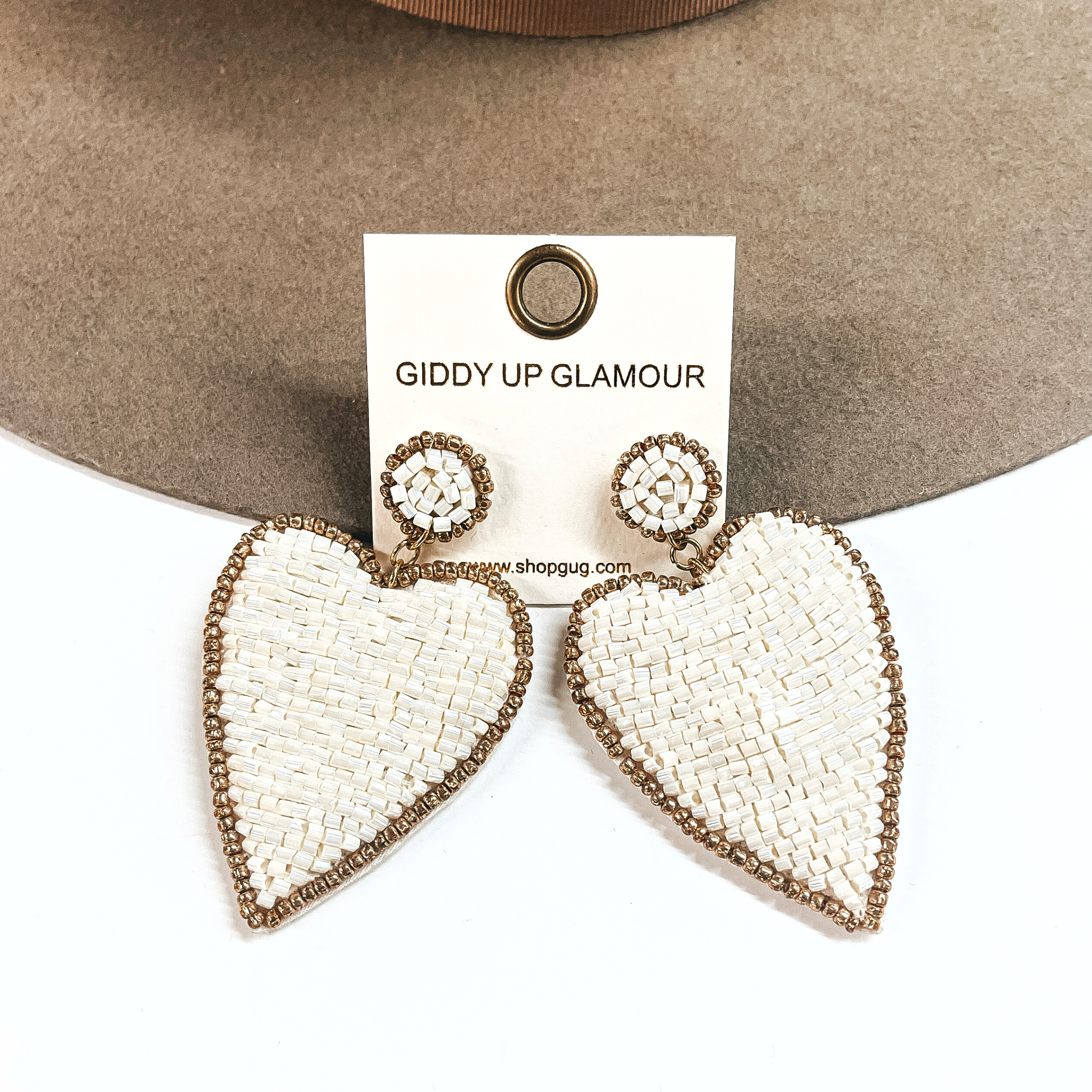These are ivory and gold beaded earrings with a heart drop. There is a gold beaded outline. These earrings are taken leaning against a light brown felt hat brim and on a white background.