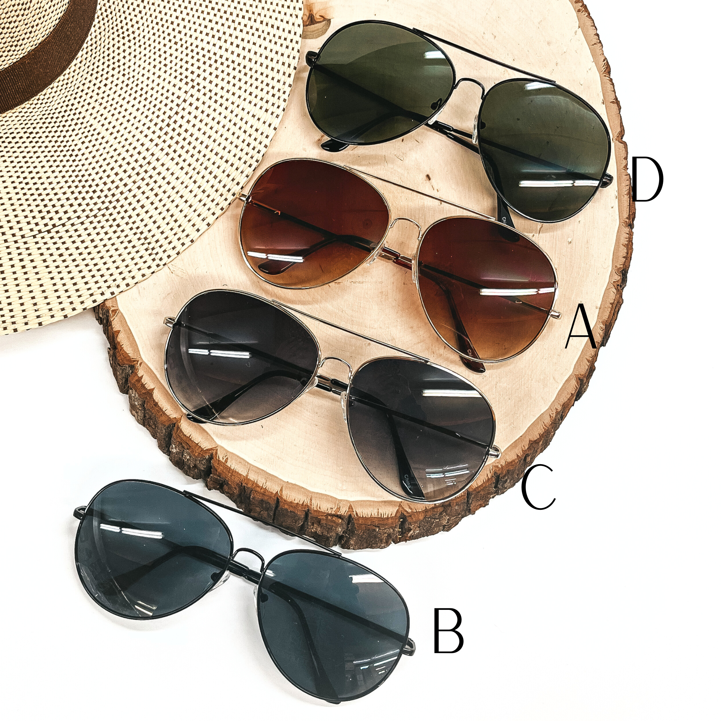 There are four pairs of aviator sunglasses in different  color frames and lenses. From top to bottom; dark green lenses with a black  frame/outline, brown lenses with a gold frame/outline, dark grey lenses  with a silver frame/outline, and black lense with a black frame/outline.  These sunglasses are taken on a slab of wood with a straw hat in the side  as decor.