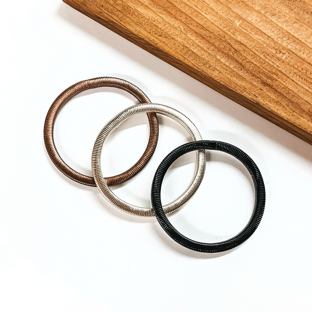 This is a set of three metallic herringone bracelets. From left to right; bronze, silver, and black. This bracelet set is taken on a white background with a slab of wood in the back as decor.