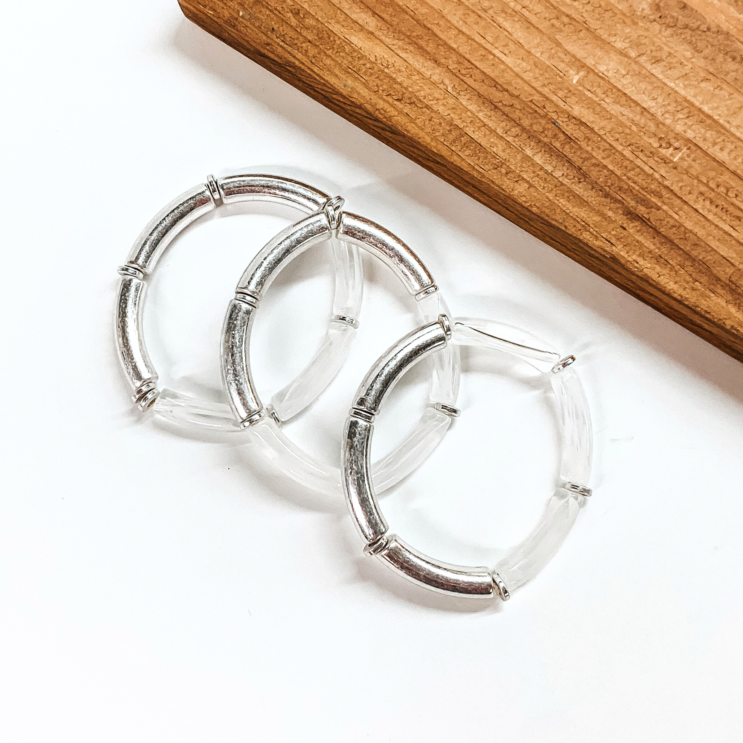This is a set of three tube bracelets in metallic silver and clear, with silver spacers. These bracelets are taken on a white background with a slab of wood in the back as decor.