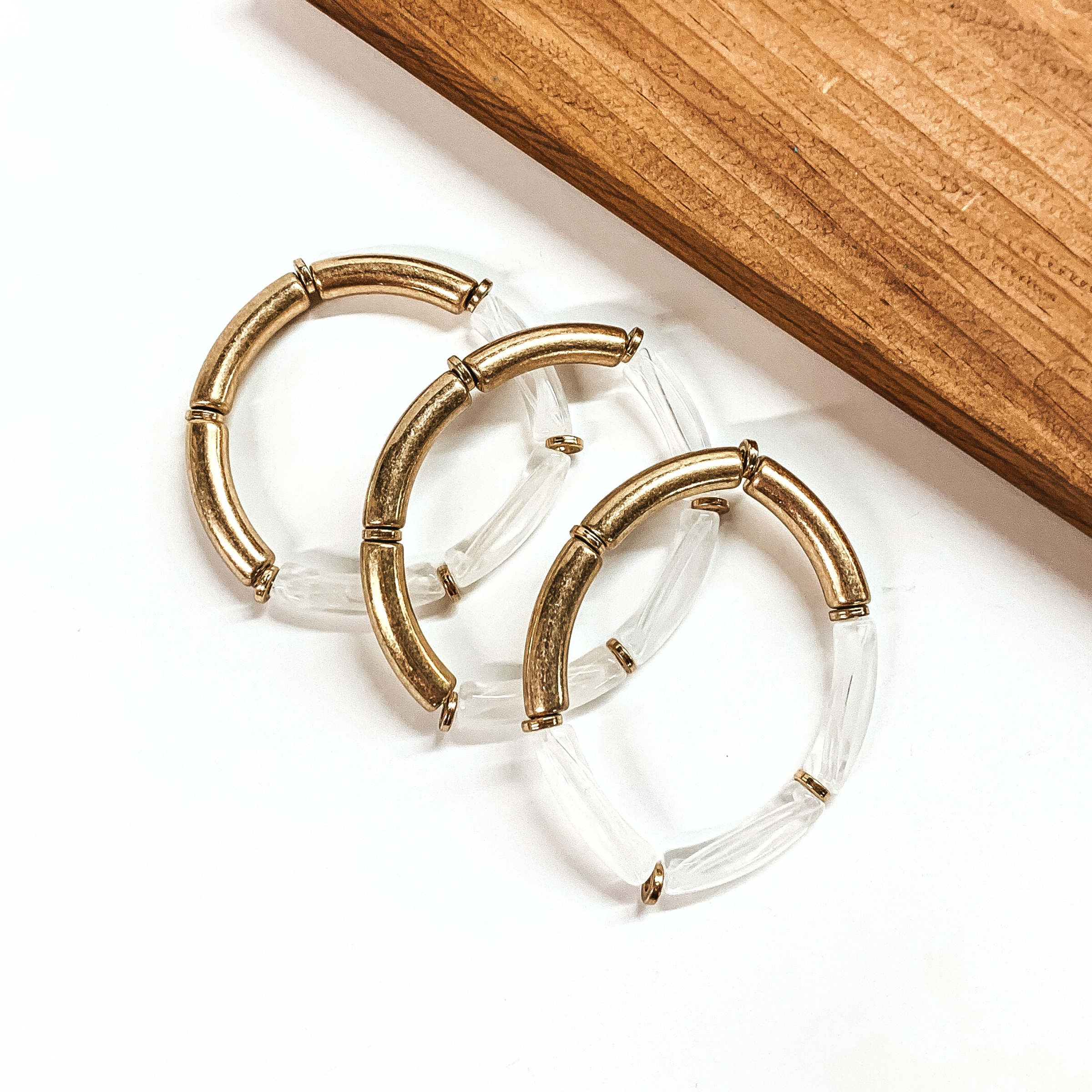 This is a set of three tube bracelets in metallic gold and clear, with gold spacers. This bracelet set is taken on a white background with a slab of wood in the back as decor.