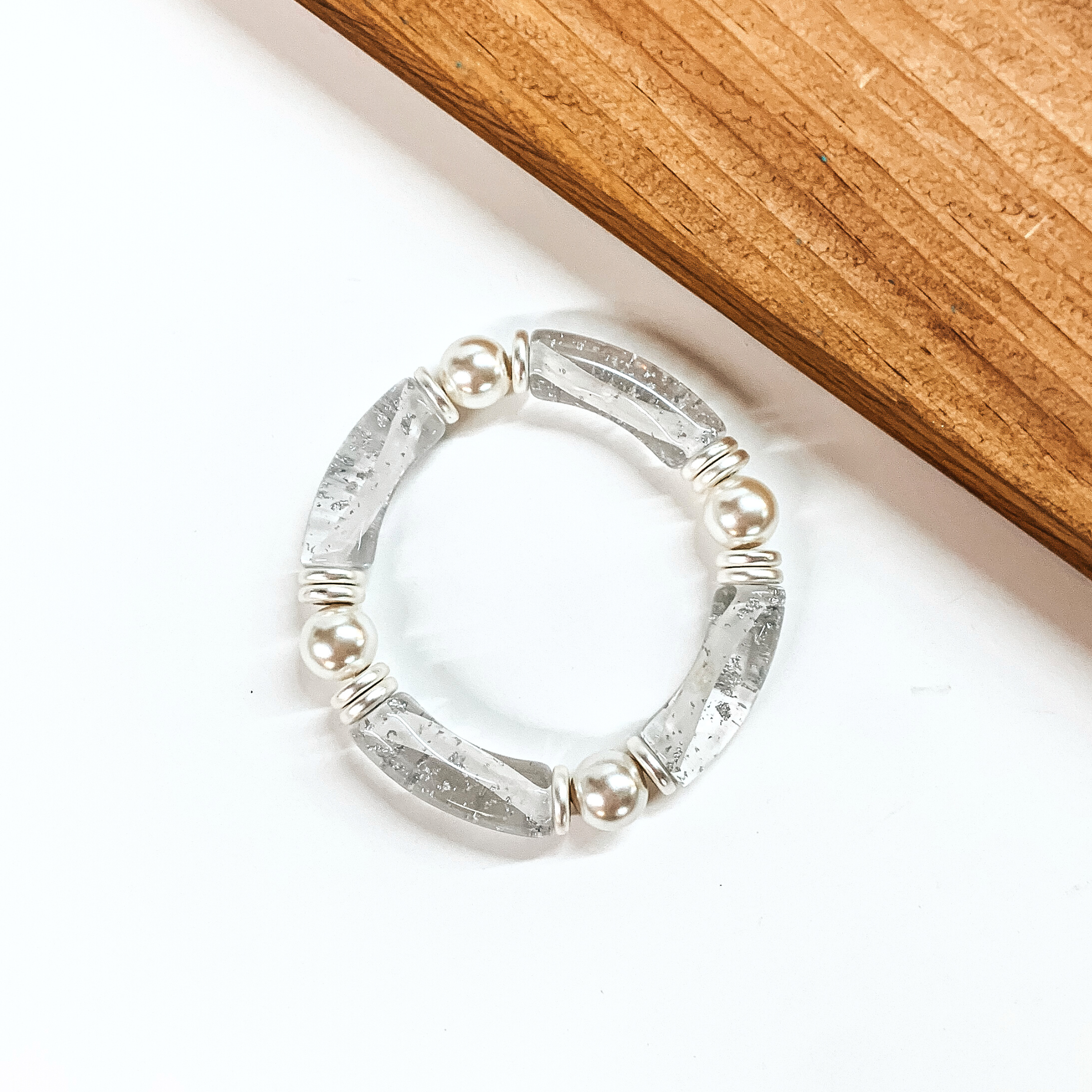 This is a clear tube bracelet with silver flakes inside. There are silver and pearl spacers. This bracelet is taken on a white background with a slab of wood in the back as decor.