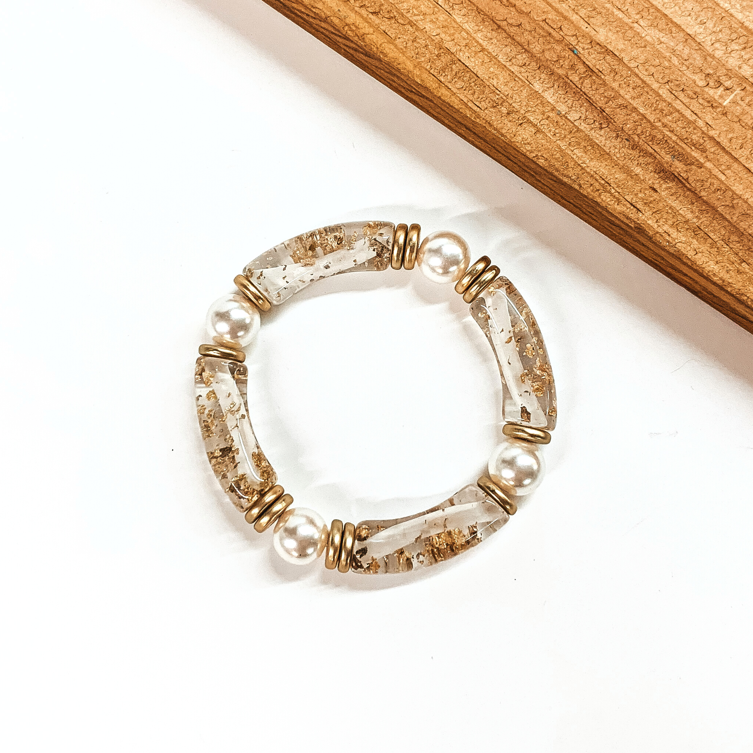 This is a clear tube bracelets with gold flakes inside and pearl spacers. This bracelet is taken on a white background with a slab of wood in the back as decor.