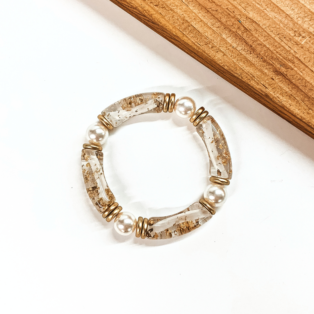 This is a clear tube bracelets with gold flakes inside and pearl spacers. This bracelet is taken on a white background with a slab of wood in the back as decor.