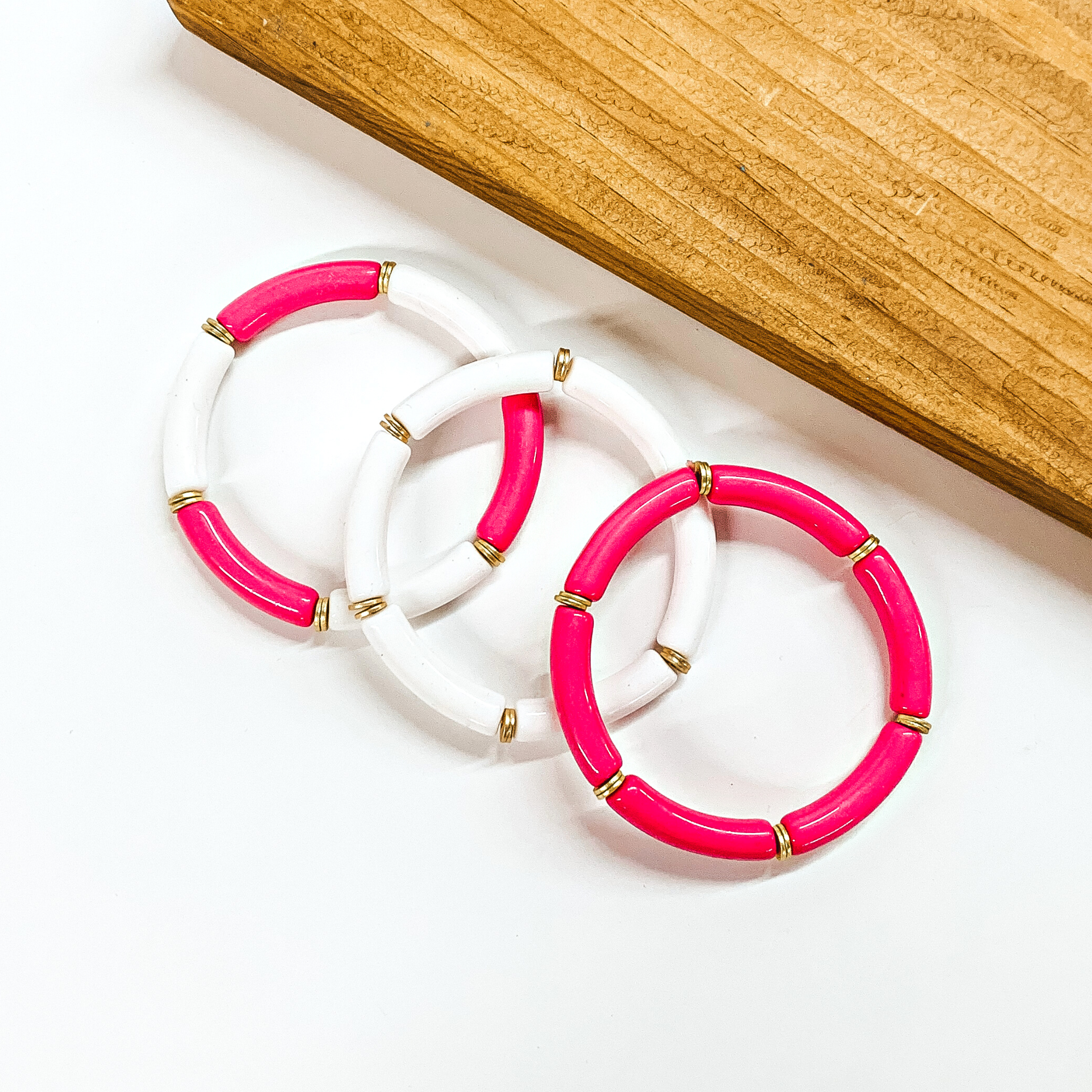 This is a set of three tube bracelets in white and hot pink with gold spacers. From left to bottom; hot pink and white tubes, white tubes, hot pink tubes. This bracelet set is taken on a white background with a slab of wood in the back as decor.