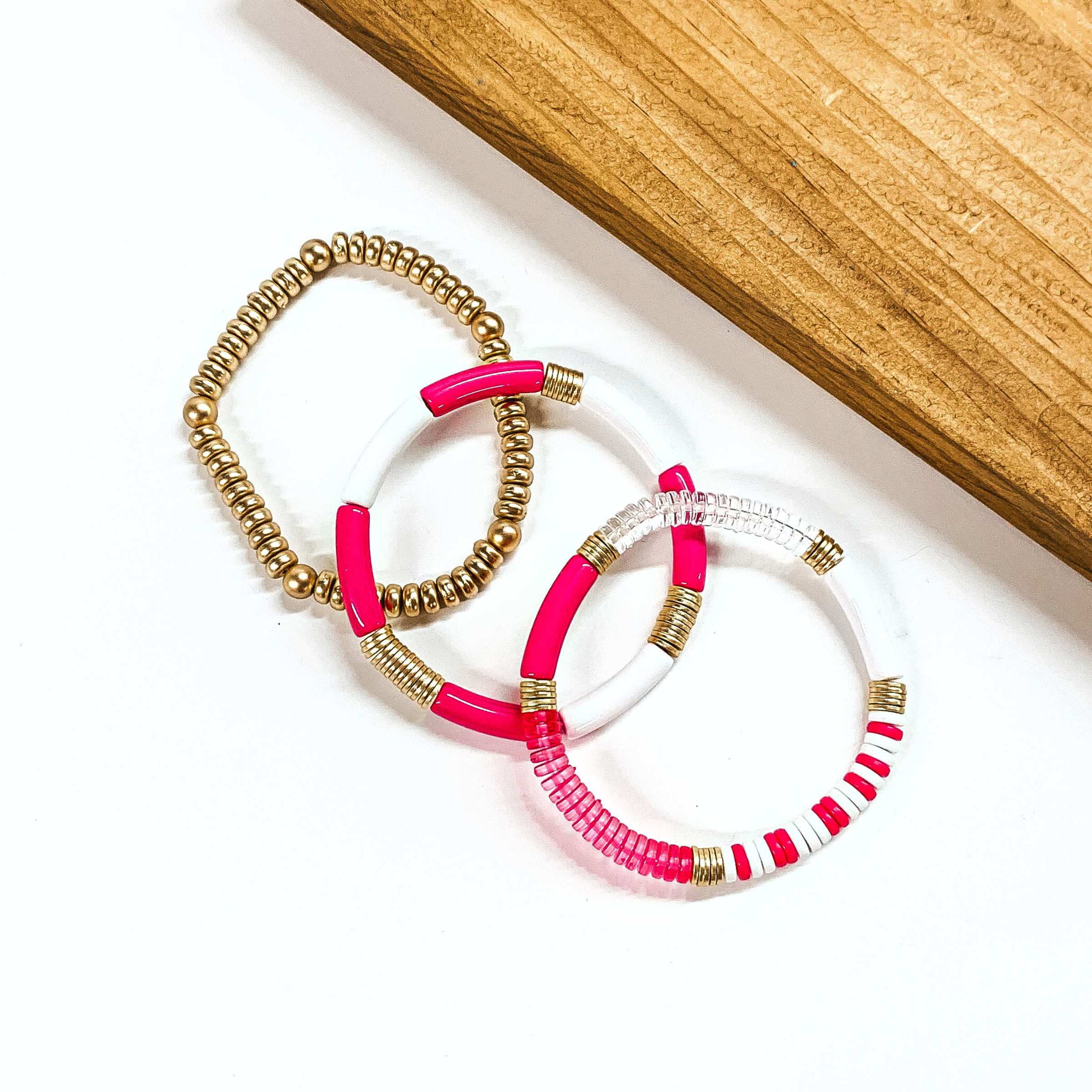 This is a set of three bracelets in hot pink, gold, and white. From left to right; gold beaded bracelet, hot pink and white tube bracelet with gold spacers. Last bracelet has hot pink and white beads in different shapes/forms with gold spacers and transparent pink and clear beads. This bracelet set is taken on a white background with a slab of wood in the back as decor.