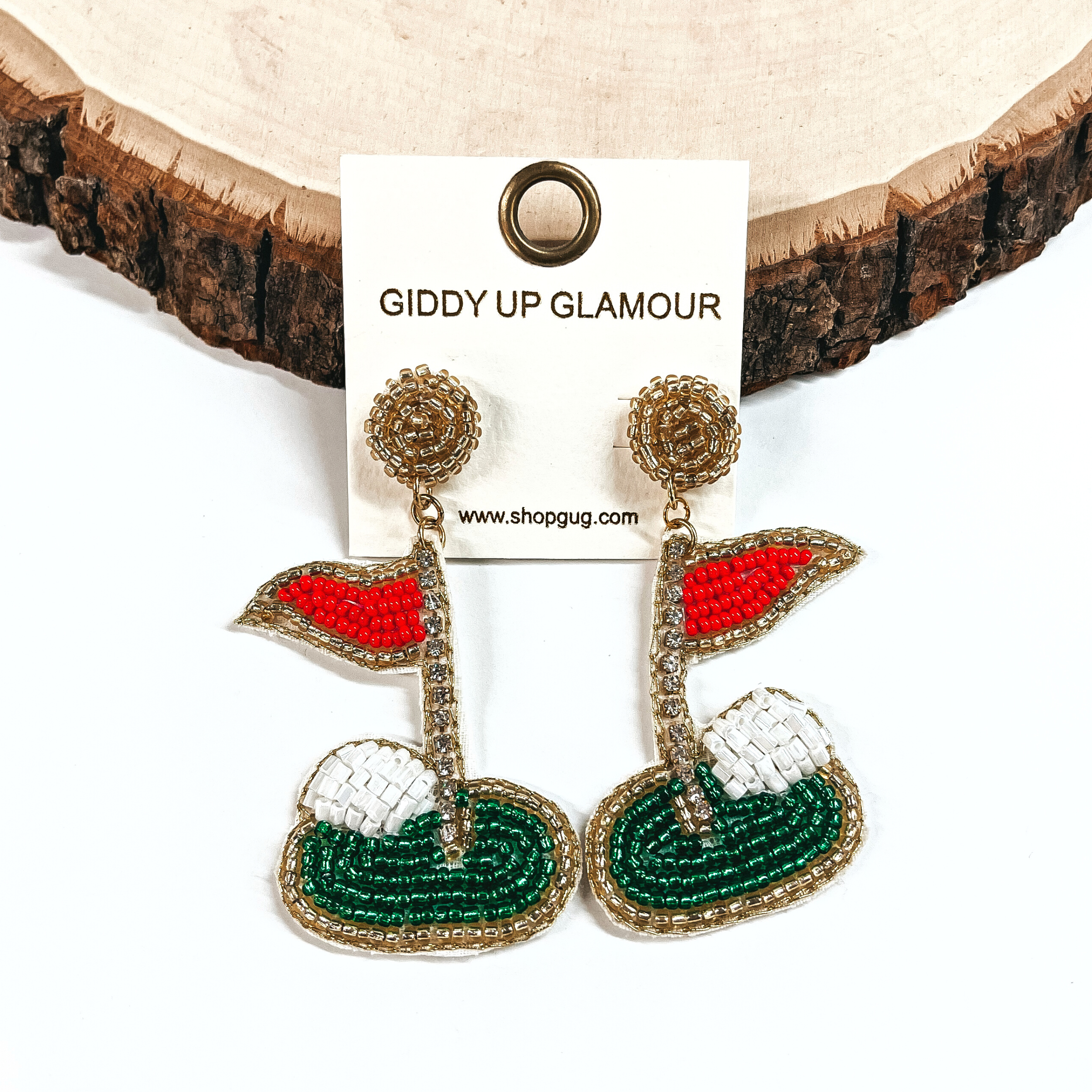 These are beaded golf earrings in mostly gold with a red flag and green grass, white ball. The flag pole has clear rhinestones. These earrings are taken on a white background and leaning against a a slab of wood.