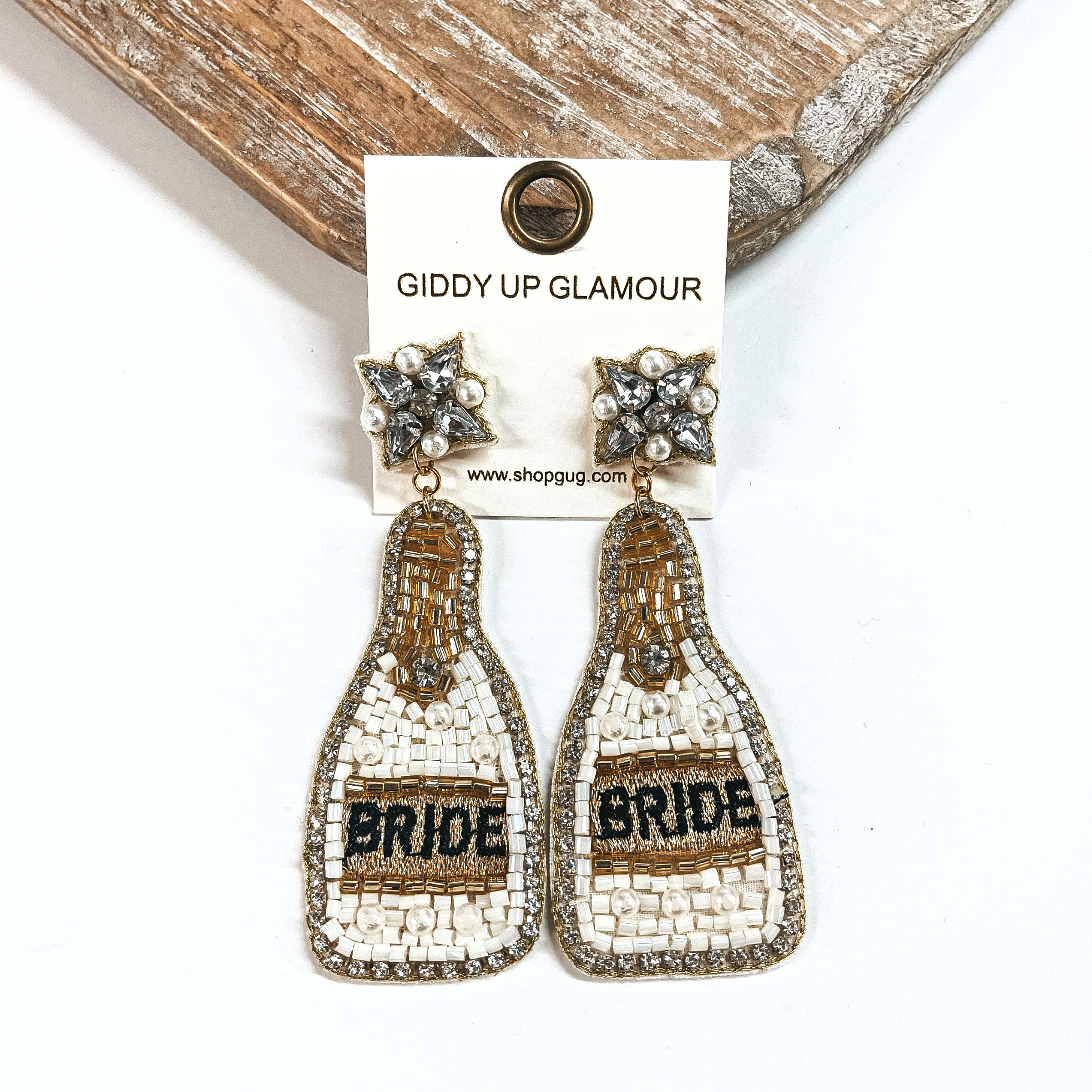 These are beaded bottle earrings in ivory mix with pearls. The postback  has clear crystals and pearls with gold stitching around. The bottle drop has  clear rhinestones all around with ivory, gold, and pearl beads. In the  center it says, Bride, in black and in a gold background. These earrings are  taken on a white background and leaning against a light wood slab.