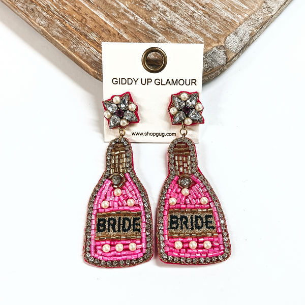 These are beaded bottle earrings in hot pink mix with pearls. The postback  has clear crystals and pearls with gold stitching around. The bottle drop has  clear rhinestones all around with hot pink, gold, and pearl beads. In the  center it says, Bride, in black and in a gold background. These earrings are  taken on a white background and leaning against a light wood slab.
