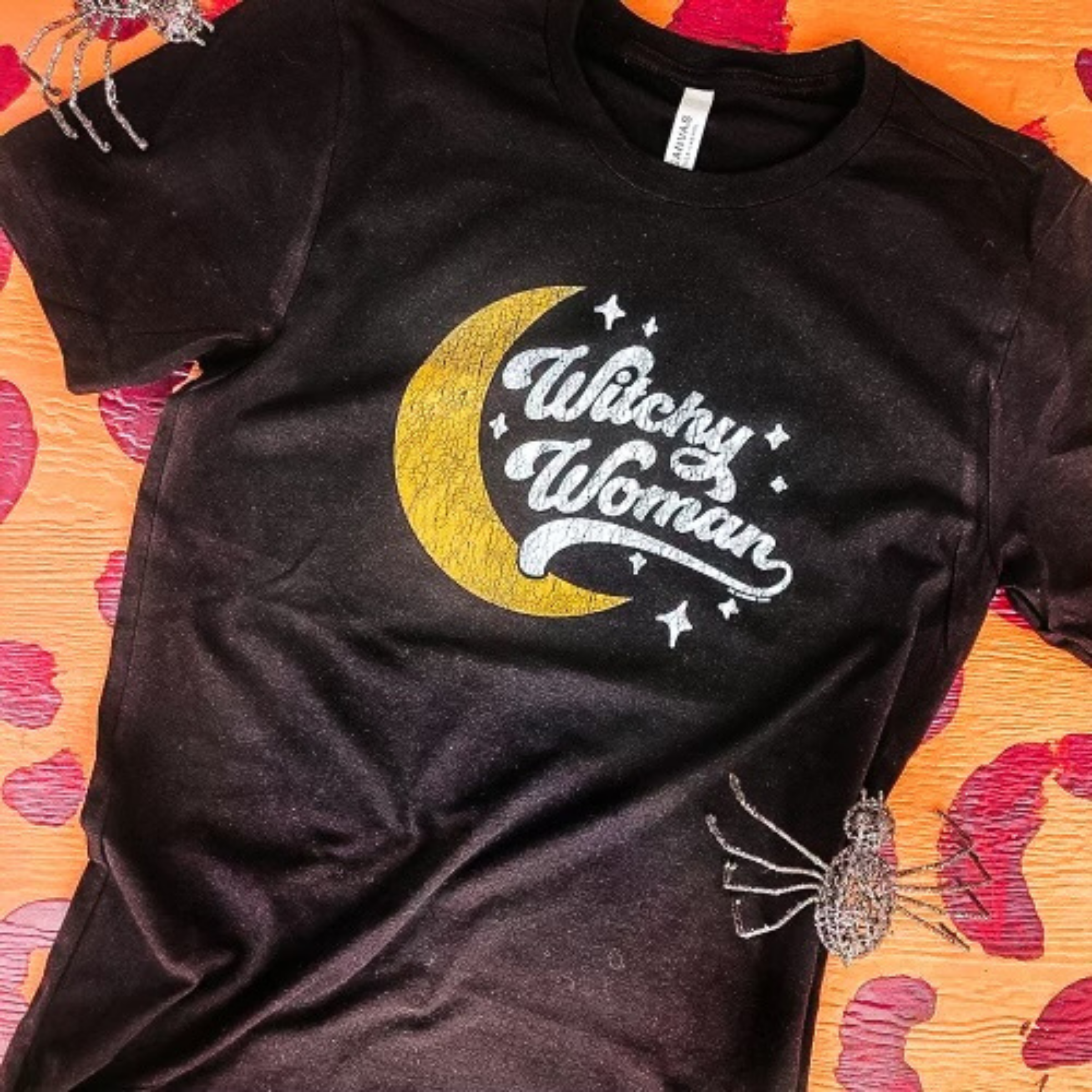 black graphic tee that reads "witchy woman" in cute font with stars and a crescent moon on shirt. Laying on a leopard print backdrop with fake spiders as props. 