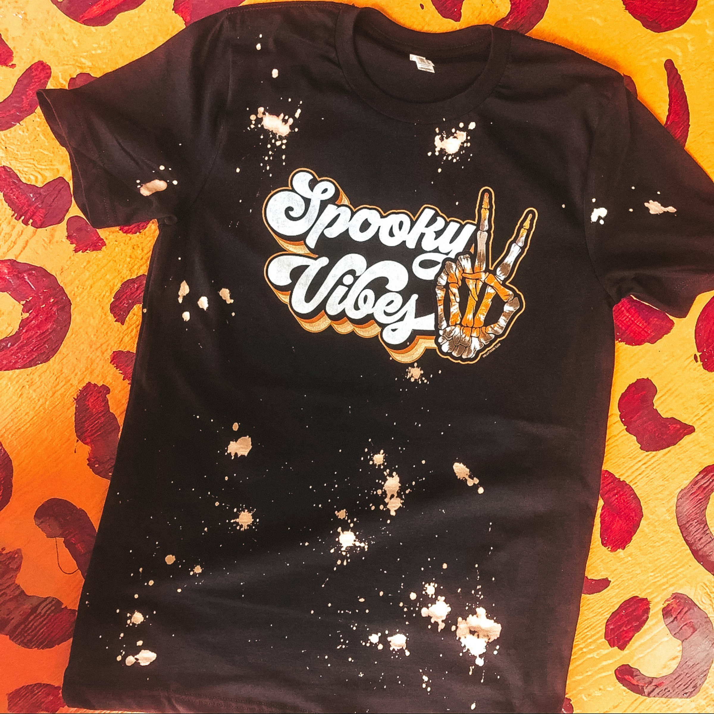 splatter graphic tee in black which reads "spooky vibes" in a groovy font with a skeleton hand doing a peace sign gesture. Tee is laying on a leopard print backdrop. 